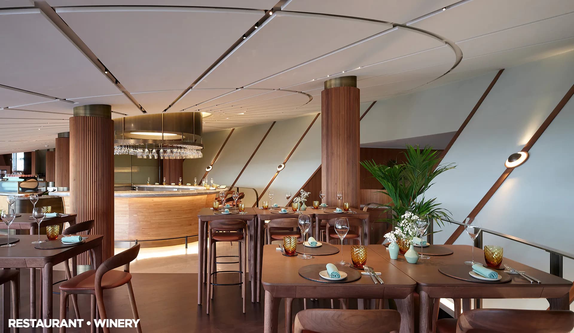 Modern restaurant and winery interior featuring curved wooden ceiling elements, fluted columns, dining tables, and a bar counter with warm lighting by Su Ergeneli.