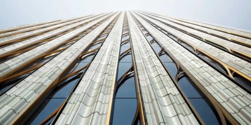 Upward perspective of a tall modernist building facade with vertical stone-clad columns and repetitive window openings against a light sky by Sue Elkattawy.