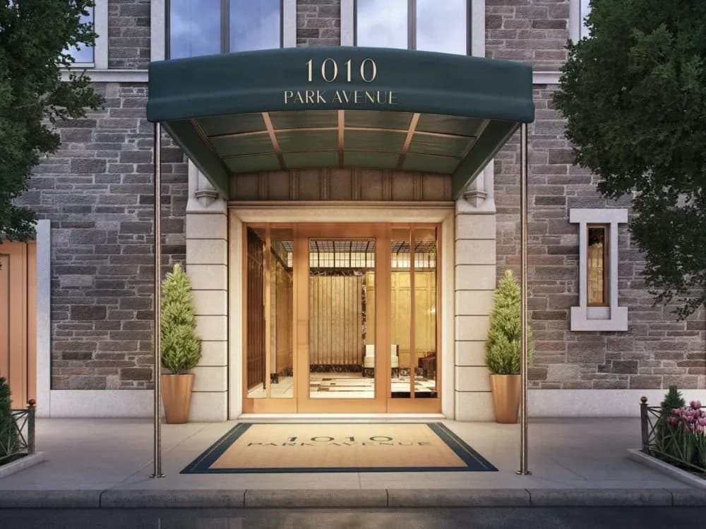 Modern luxury residential building entrance at 1010 Park Avenue featuring a dark green canopy, brass-framed glass doors, and warm interior lighting, by Sue Elkattawy.