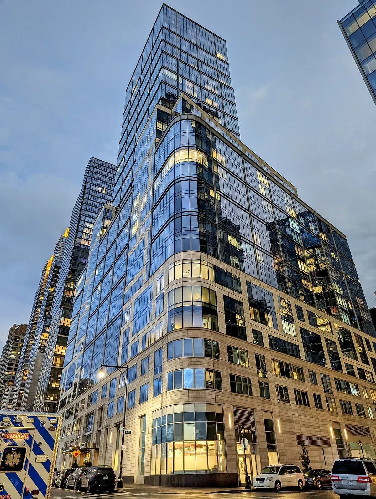 Modern curved glass and stone office tower with illuminated interiors at dusk, featuring distinctive rounded corner bay windows and mixed material facade by Sue Elkattawy.