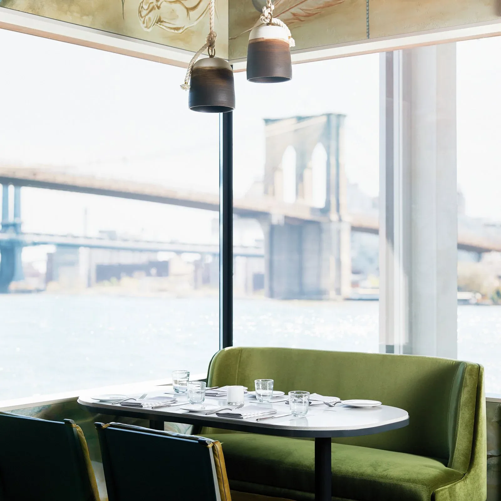 Modern dining nook with olive-green banquette seating, black pendant lights, and floor-to-ceiling windows overlooking a city waterfront by Sue Elkattawy.