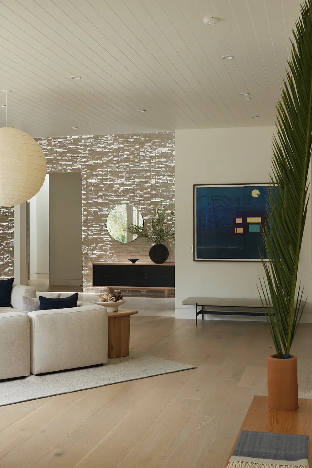 Modern living room with cream upholstered seating, light wood flooring, textured stone accent wall, round mirror, and tall potted plant by Suha Samara.