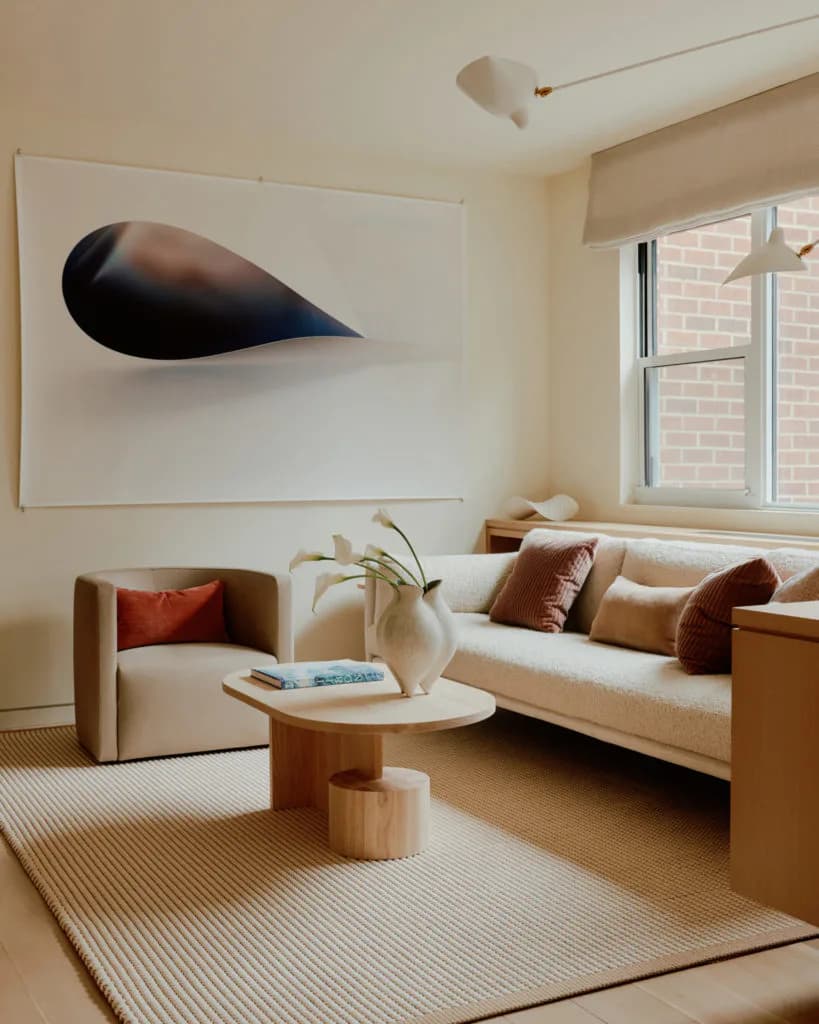 Minimalist living room with cream sofa, beige armchair, wooden side table, and abstract black teardrop artwork by Suha Samara.