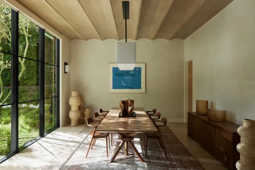 Minimalist dining room with wooden table and chairs, concrete ceiling beams, large black-framed glass doors overlooking greenery, and blue artwork, designed by Suha Samara.