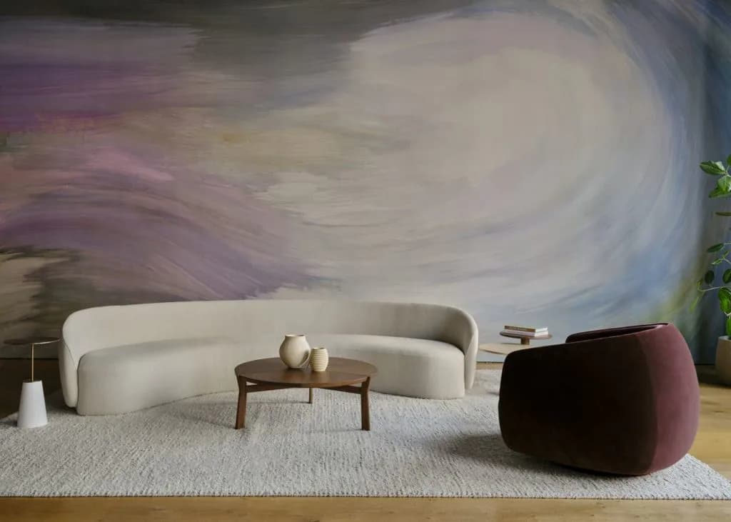 Modern living room with curved cream sofa, wooden side table, burgundy pouf, and abstract swirling mural backdrop in soft purples and whites by Suha Samara.