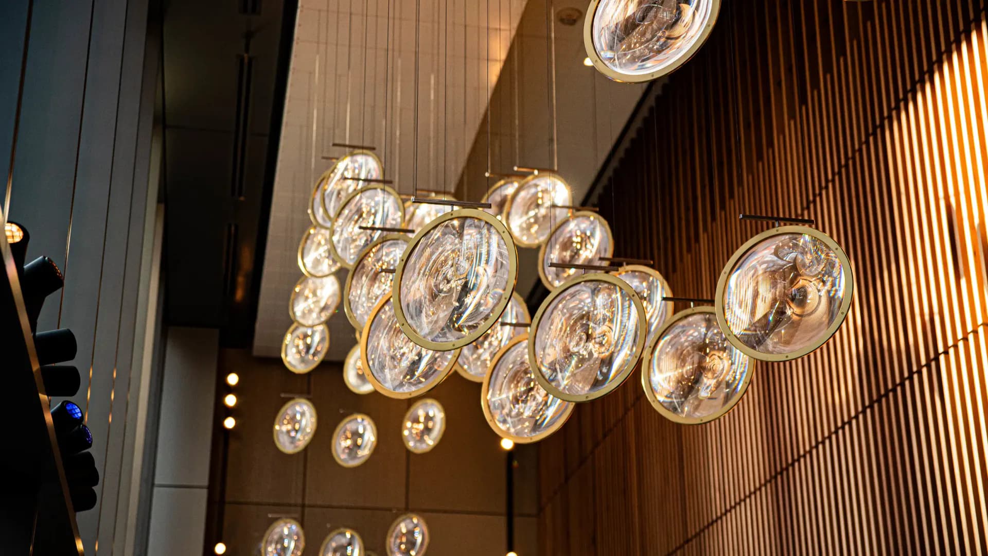 Modern ceiling installation featuring cascading spherical glass pendant lights with warm illumination against wooden slat walls by Sunmi Lee.