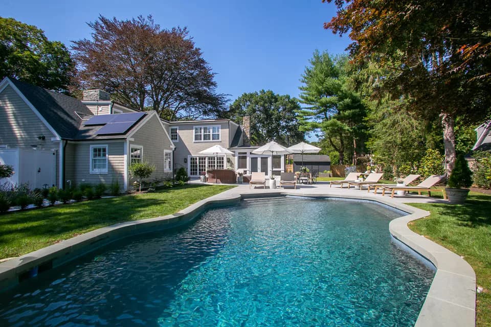 Luxurious residential backyard featuring a curved swimming pool with turquoise water, mature landscaping, and a multi-level home with modern updates by Susan Anderson.