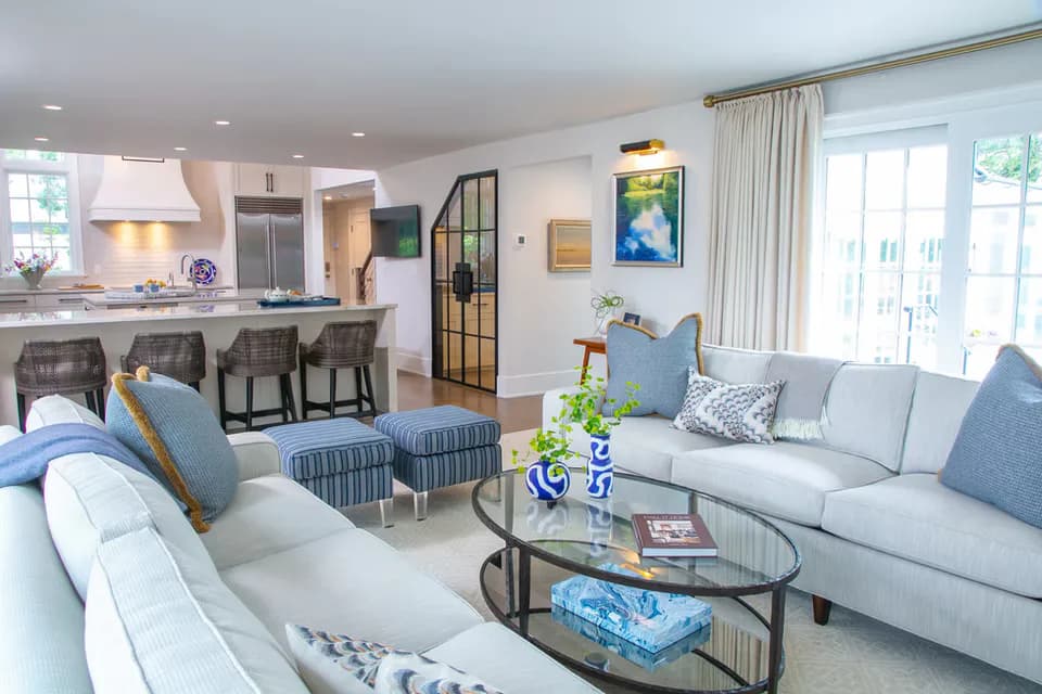 Bright coastal living room with light gray sofas, blue accent pillows, glass coffee table, and adjacent kitchen with island seating, designed by Susan Anderson.