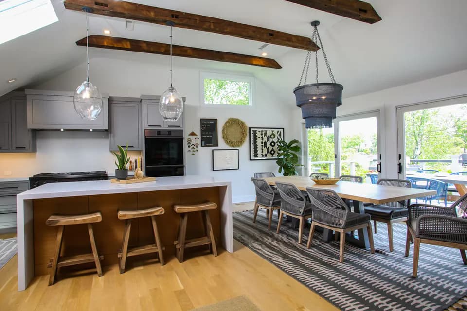 Modern open-concept kitchen and dining area featuring exposed wooden ceiling beams, white cabinetry, wooden bar stools, and a dining table with black chairs on a patterned rug, designed by Susan Anderson.