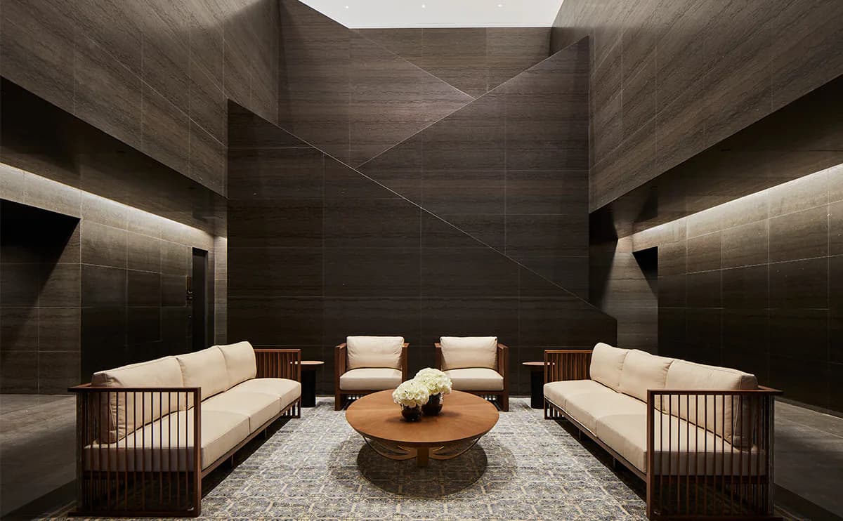 Symmetrical lounge with dark textured walls, wooden-frame seating with cream cushions, round wooden table, and skylight, designed by Susan Brady.