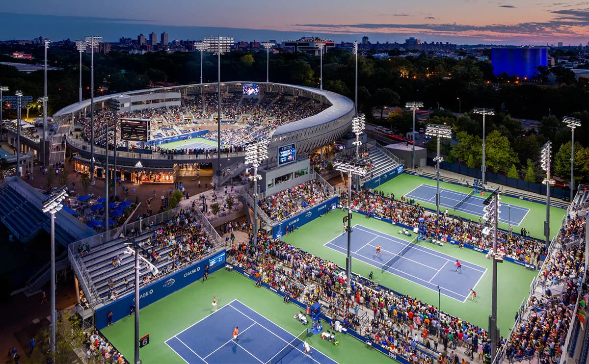 Aerial view of a modern sports complex with illuminated tennis courts and a large oval stadium at dusk, designed by Susan Brady.