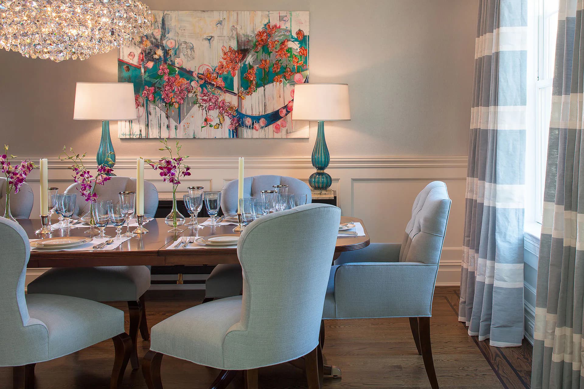 Elegant dining room with light blue upholstered chairs, wood table, teal accents, floral artwork, and striped curtains by Susan Carlson.