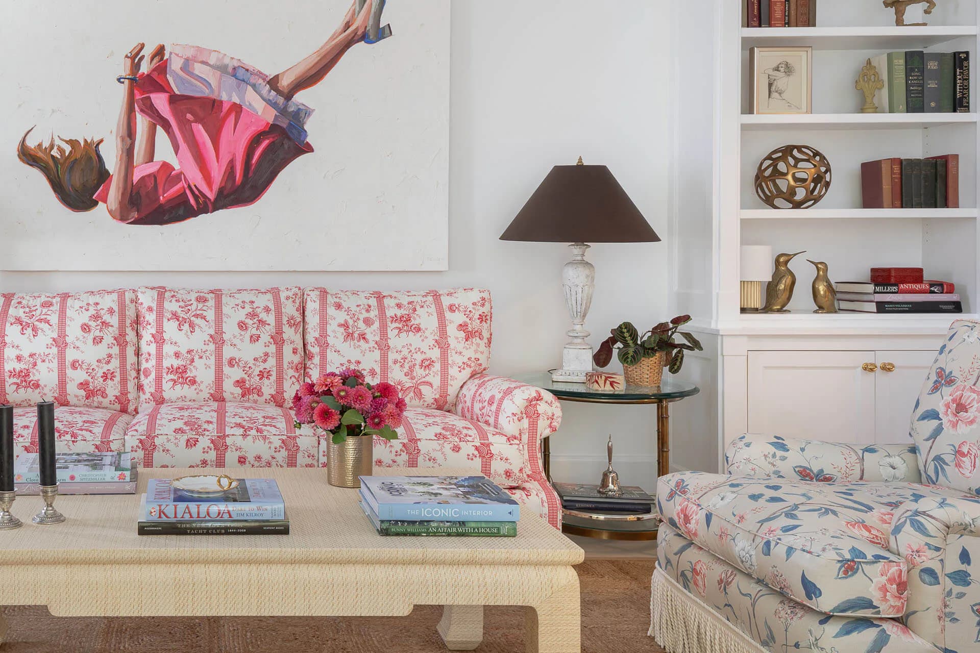 A feminine bedroom interior with pink and white toile patterned upholstered sofa and bedding, cream wood furniture, built-in shelving, and artwork by Susan Carlson.
