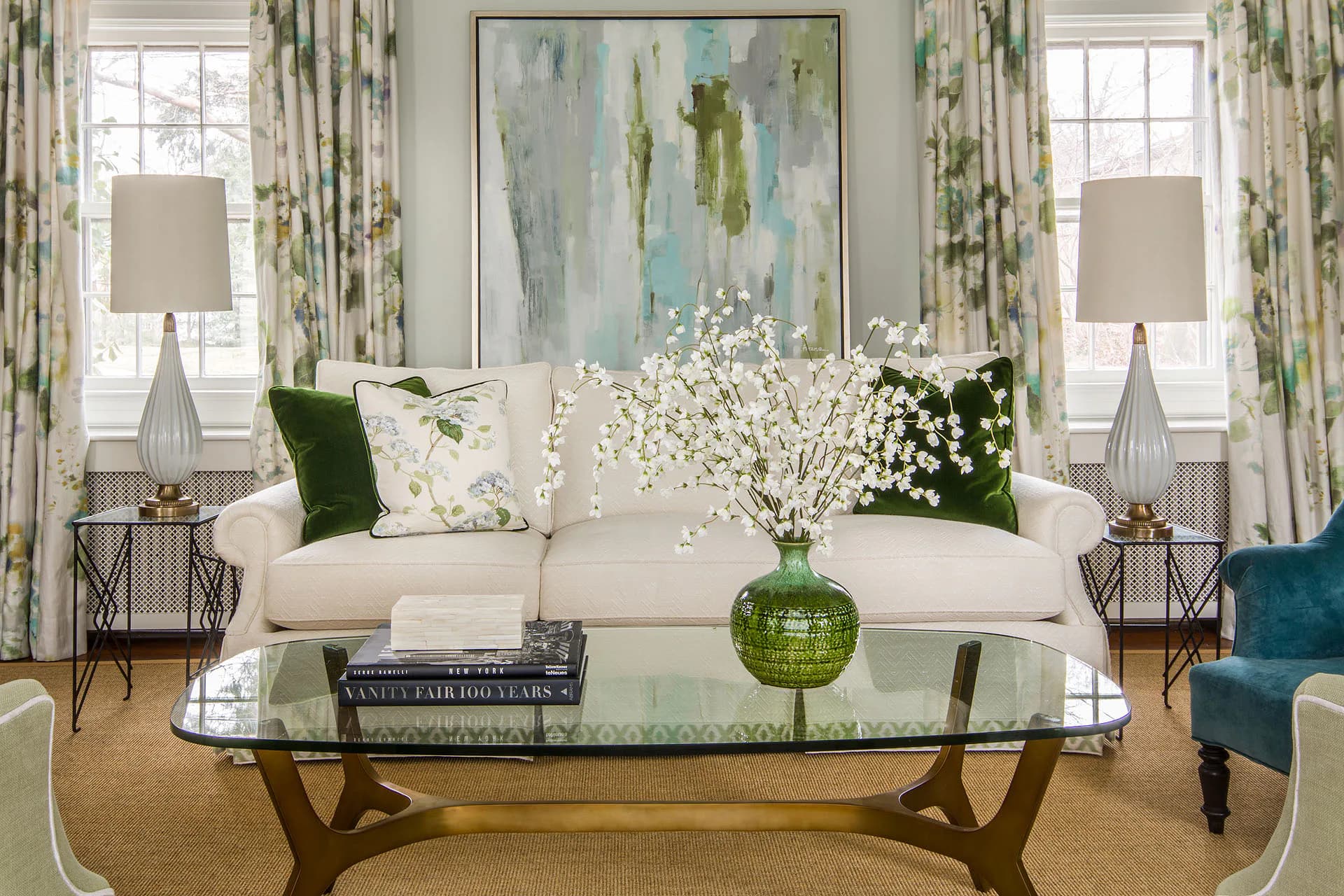 Elegant living room with cream sofa, glass coffee table, green floral arrangements, patterned curtains, and abstract artwork by Susan Carlson.
