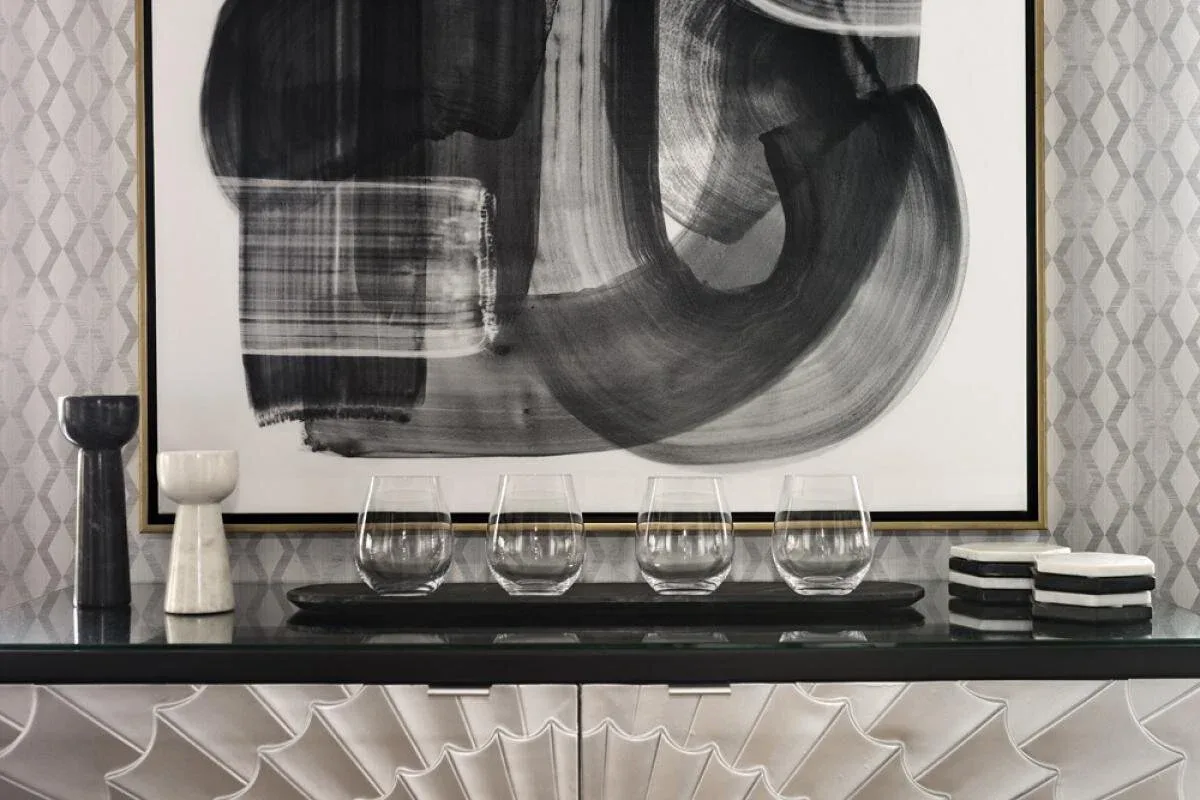 Modern black sideboard with geometric front panels displays abstract framed artwork and clear glassware against patterned wallpaper, designed by Susan Hughes.