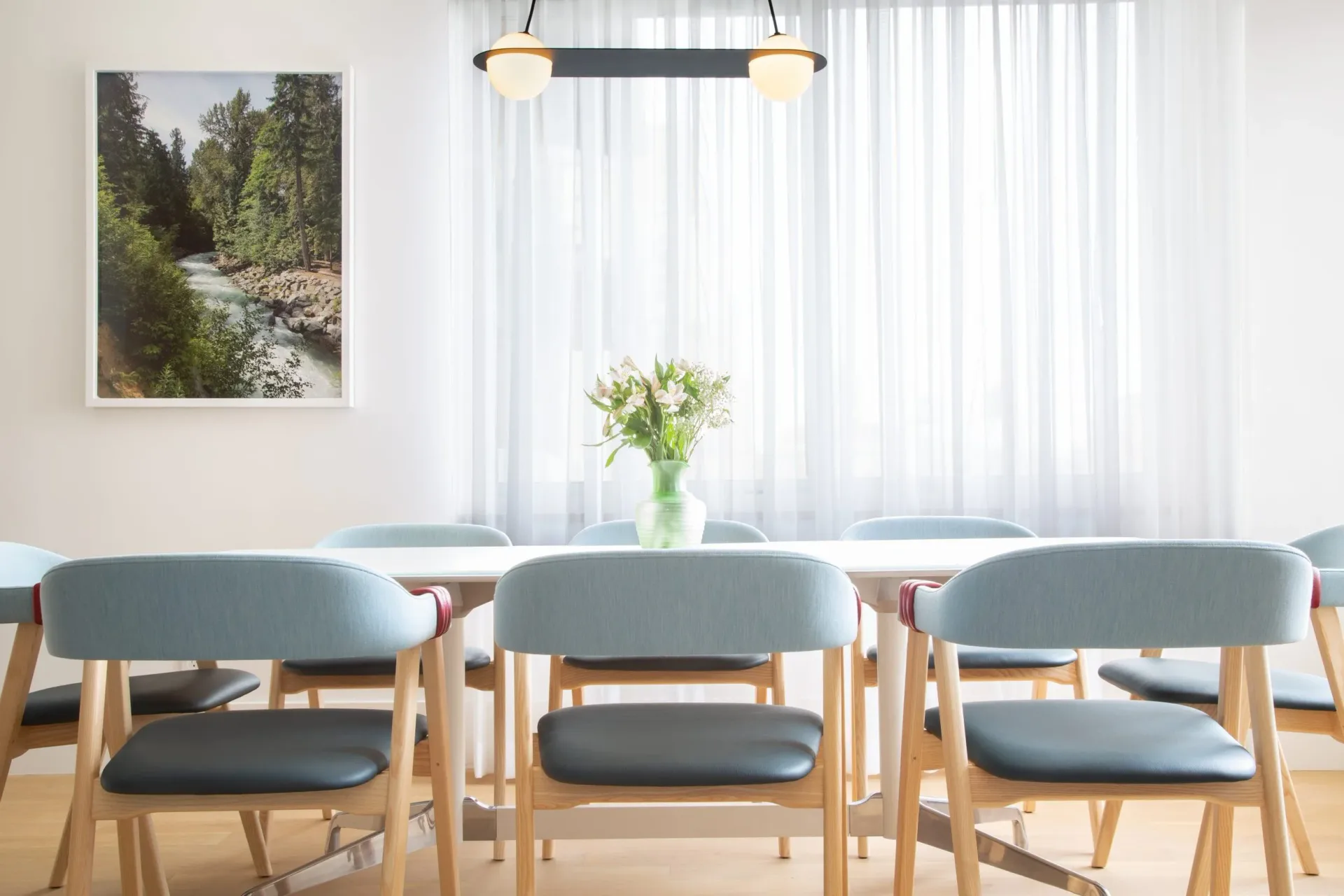Minimalist dining room with three light blue upholstered chairs, natural wood frames, and a glass vase centerpiece by Susan Torenli.