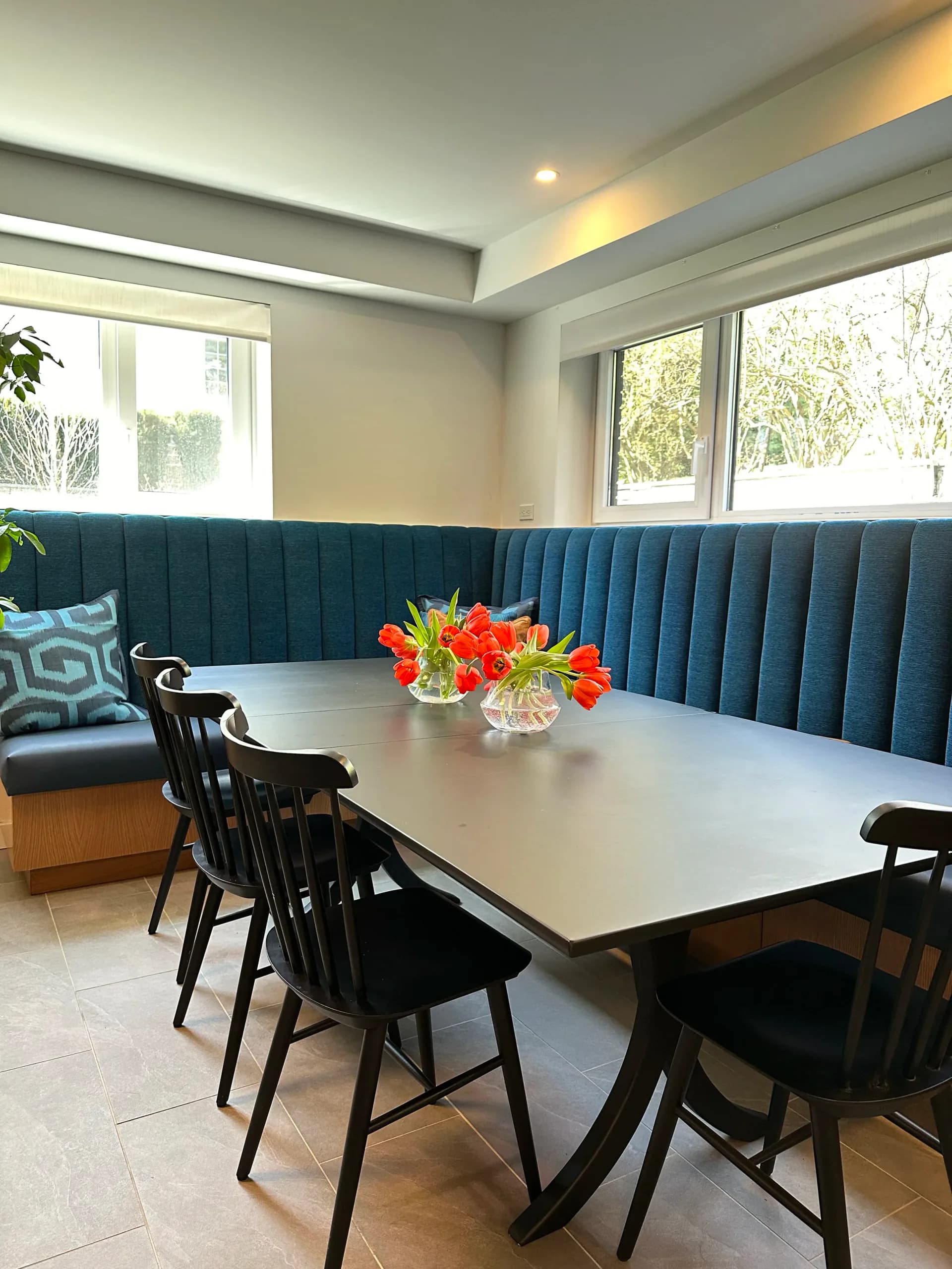 Modern dining room with teal vertical-ribbed banquette seating, white table, black wooden chairs, bright windows, and fresh orange floral arrangement by Susan Tully.