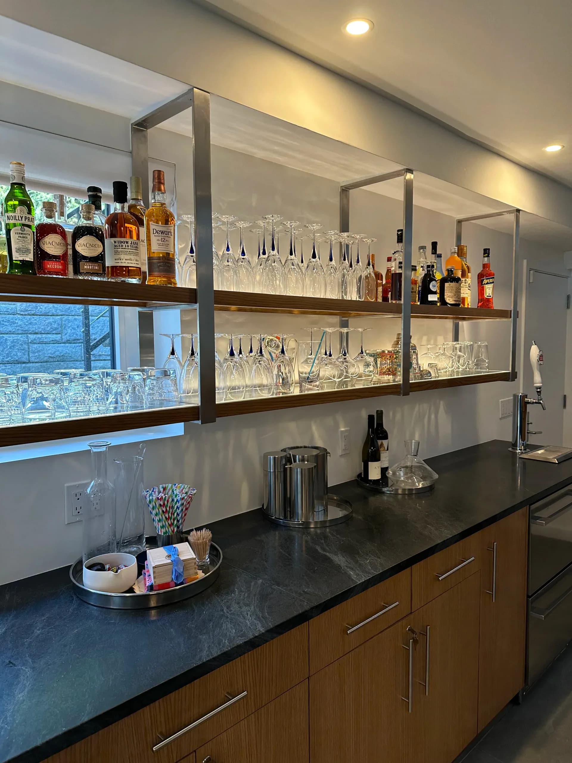 Modern home bar with floating glass shelving, dark countertop, wood cabinetry, and extensive bottle and glassware display, designed by Susan Tully.