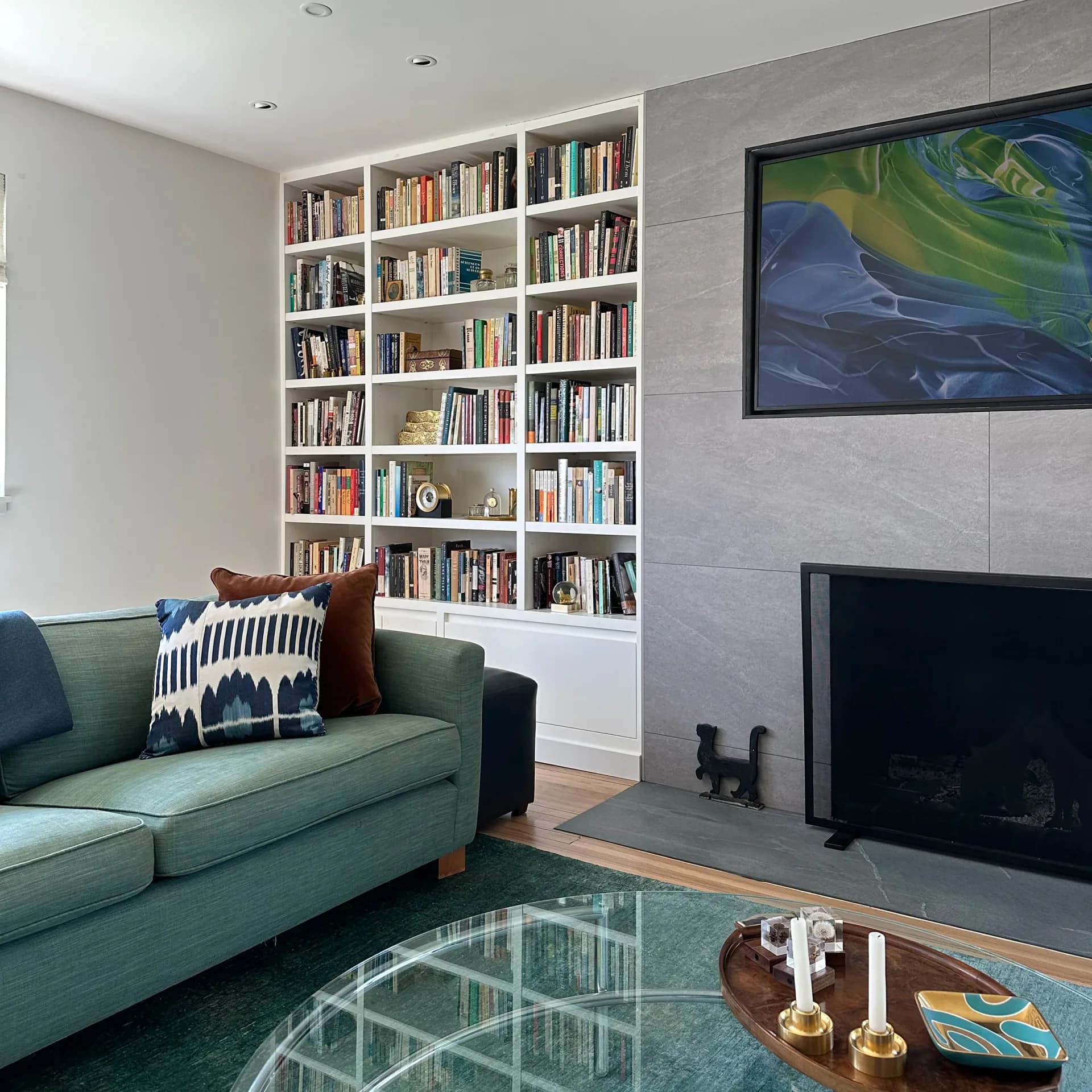 Modern living room featuring a sage green sofa, built-in floor-to-ceiling white shelving with books, fireplace with gray tile surround, TV mounted above, geometric rug, and abstract artwork by Susan Tully.