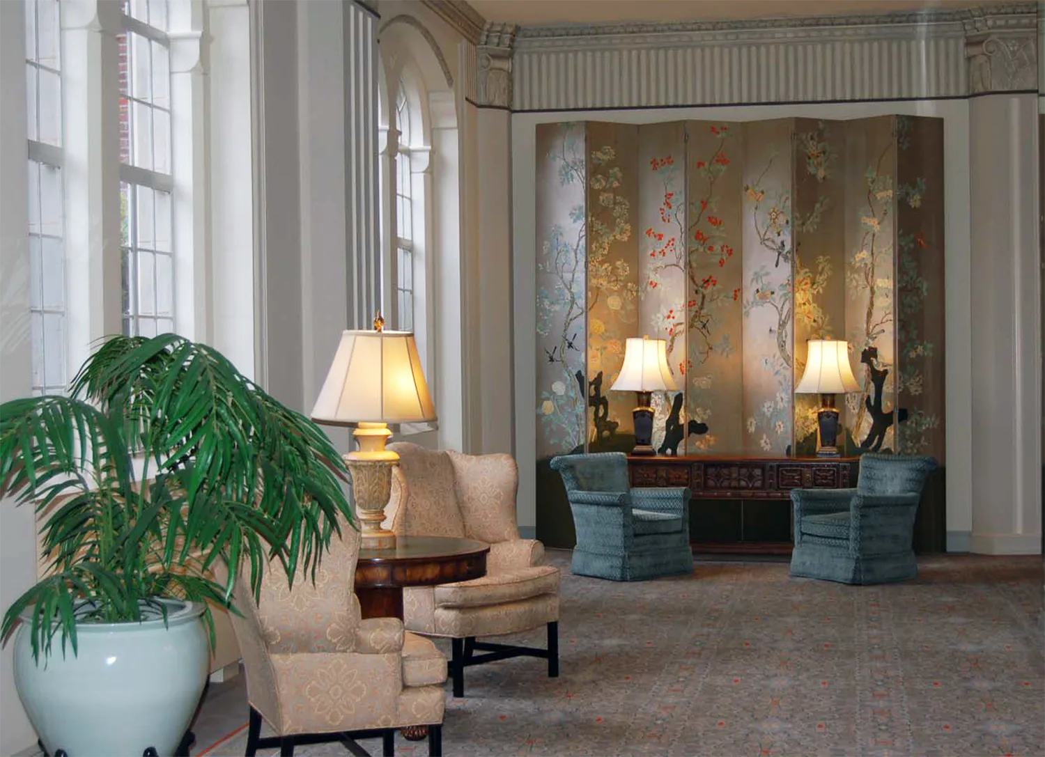 Elegant hotel lobby with beige seating, mirrored wall panels with gold leaf detailing, table lamps, and potted greenery by Susan Tully.