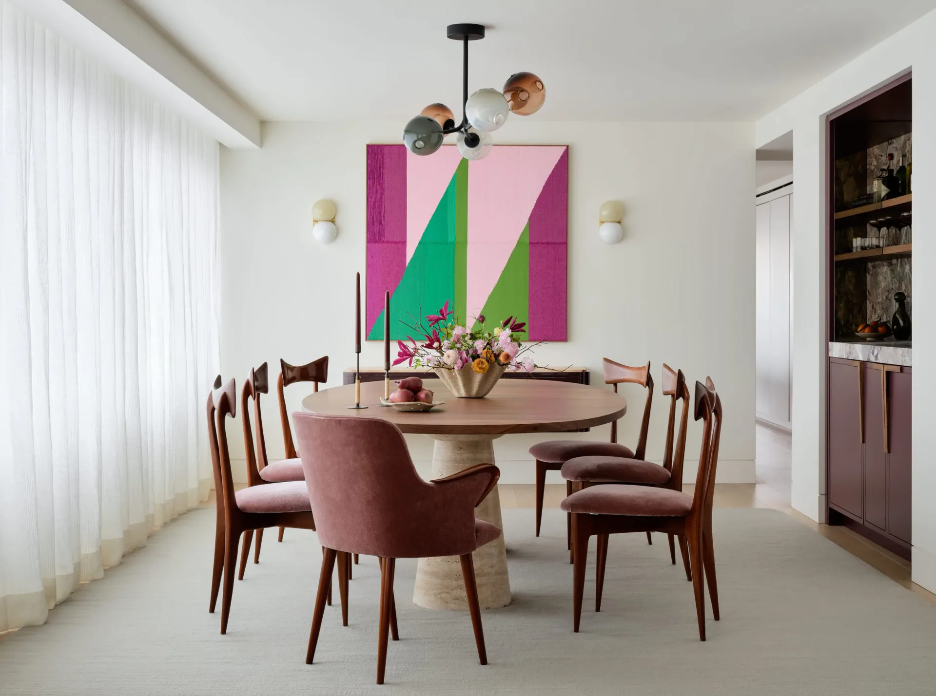 A modern dining room with a round wooden table, burgundy chairs, and geometric pink and green artwork on the white wall, designed by Susana Simonpietri.