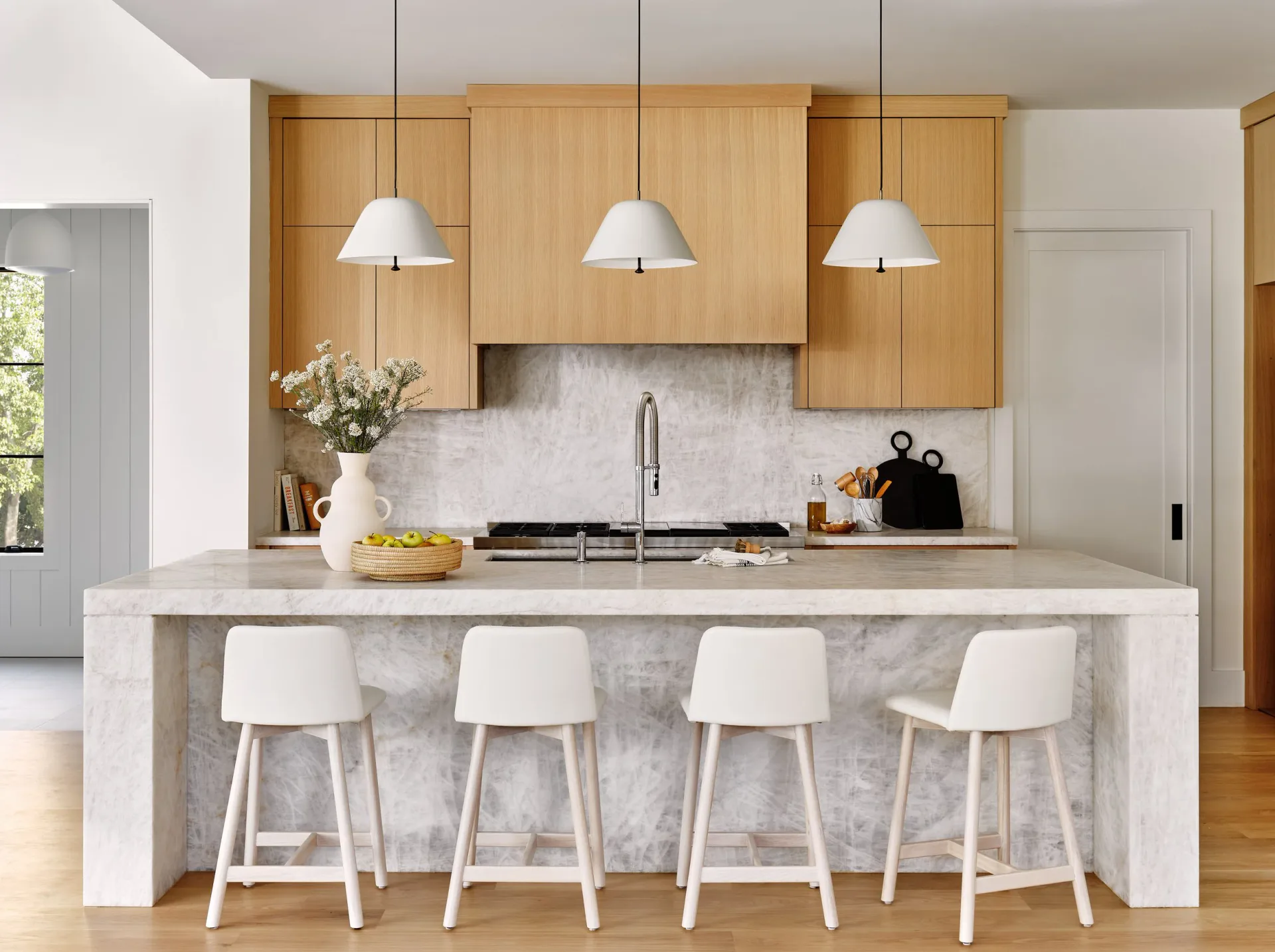 Modern kitchen with natural wood cabinetry, white marble waterfall island, pendant lighting, and white bar stools in a bright, minimalist space by Susana Simonpietri.