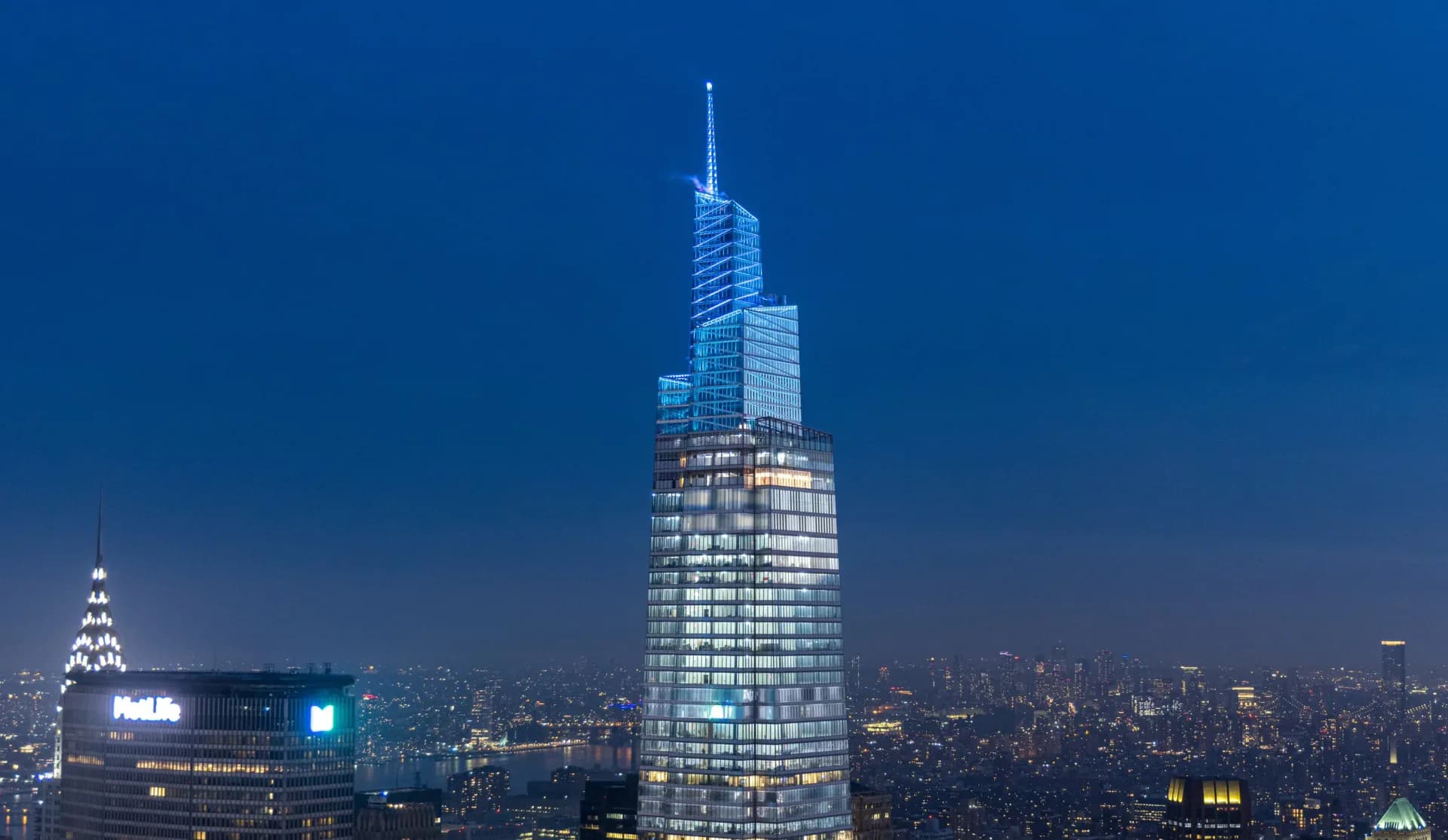 Illuminated skyscraper tower with stepped glass facade rising above city skyline at dusk by Suzan Tillotson.
