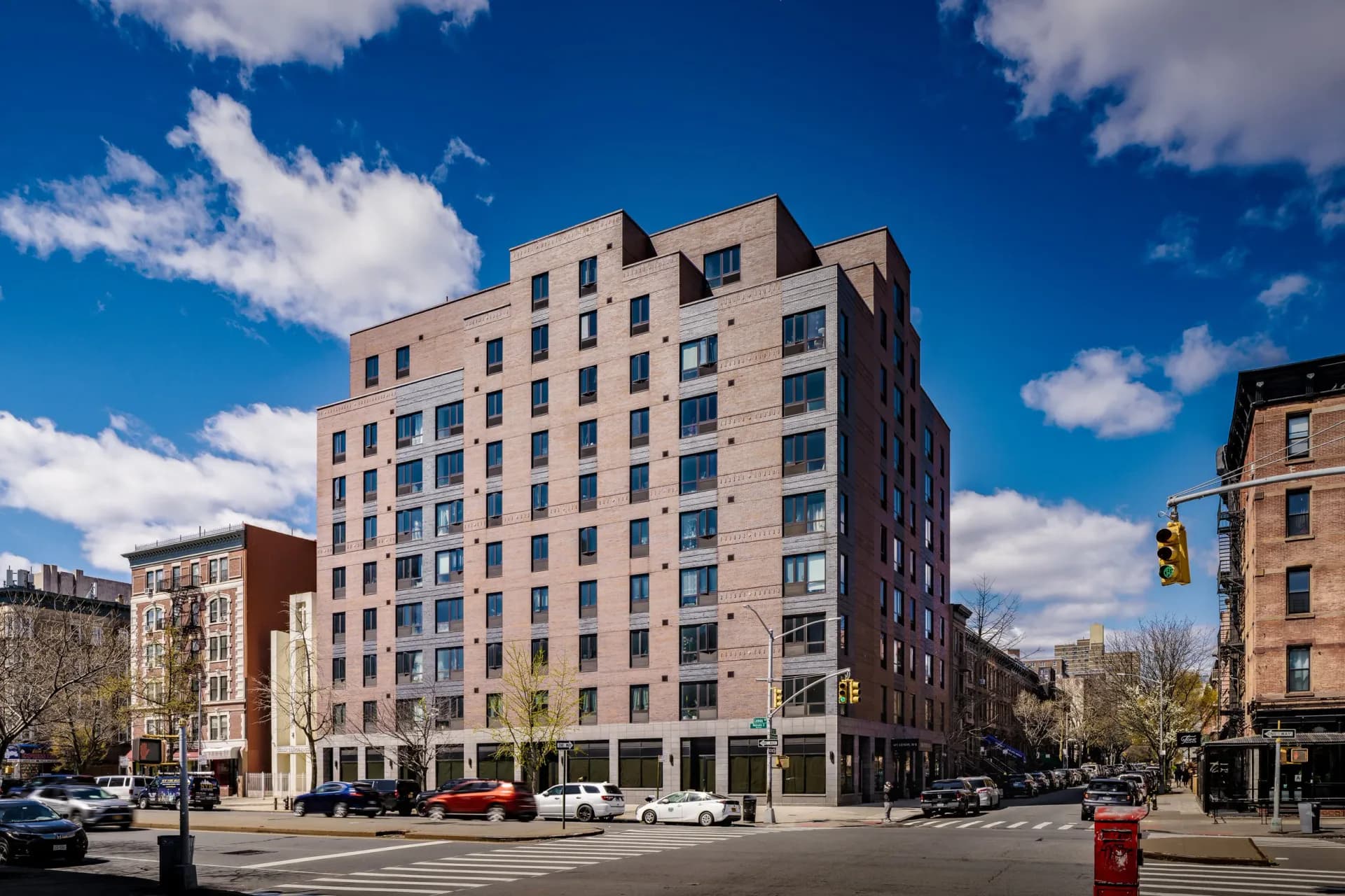 Contemporary residential building with tan brick facade and regular window grid overlooking an urban intersection with blue sky by Suzanna Tharian.