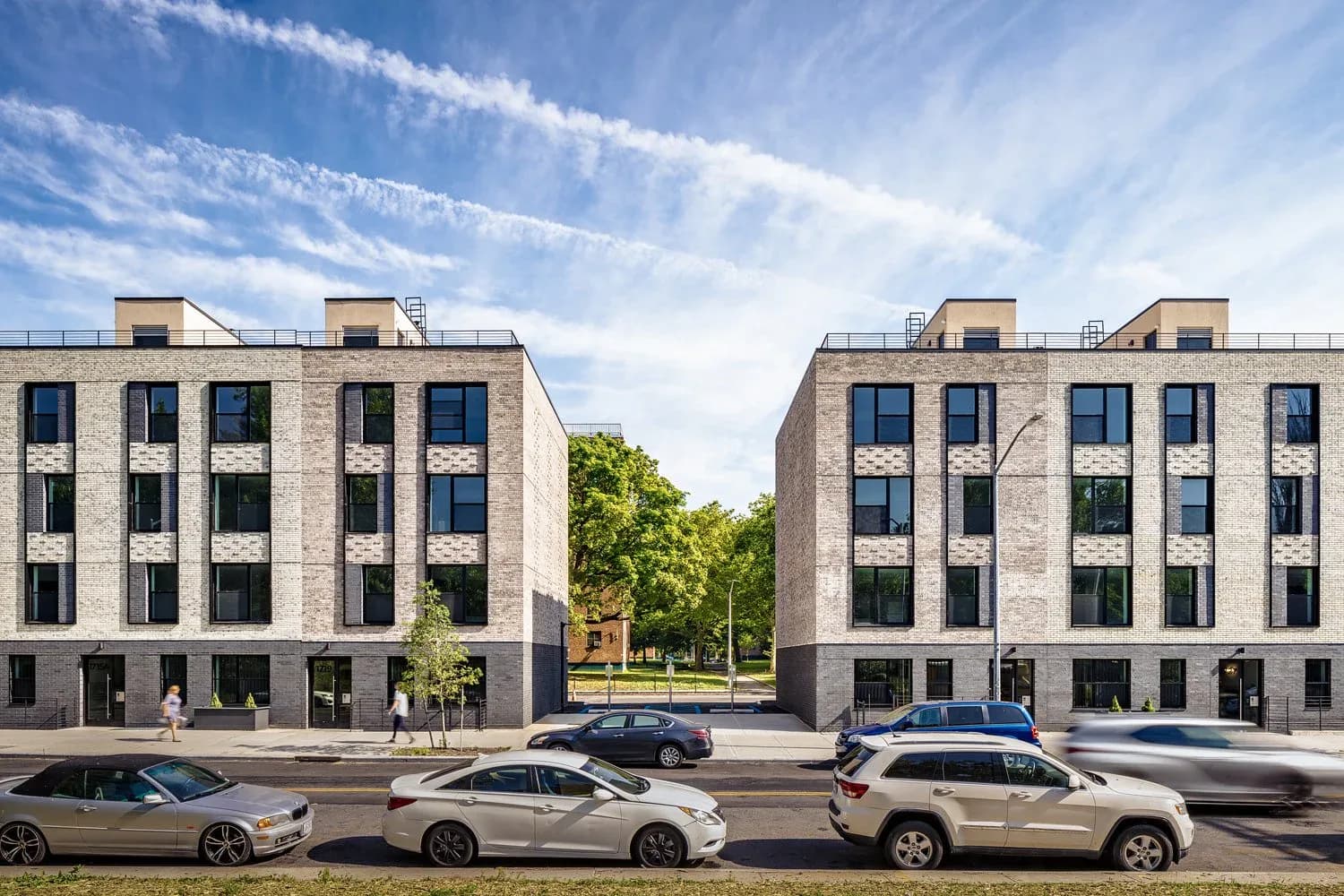 Contemporary residential building facade with cream-colored brick, large windows, and ground-floor retail, fronted by parked cars and green trees by Suzanna Tharian.