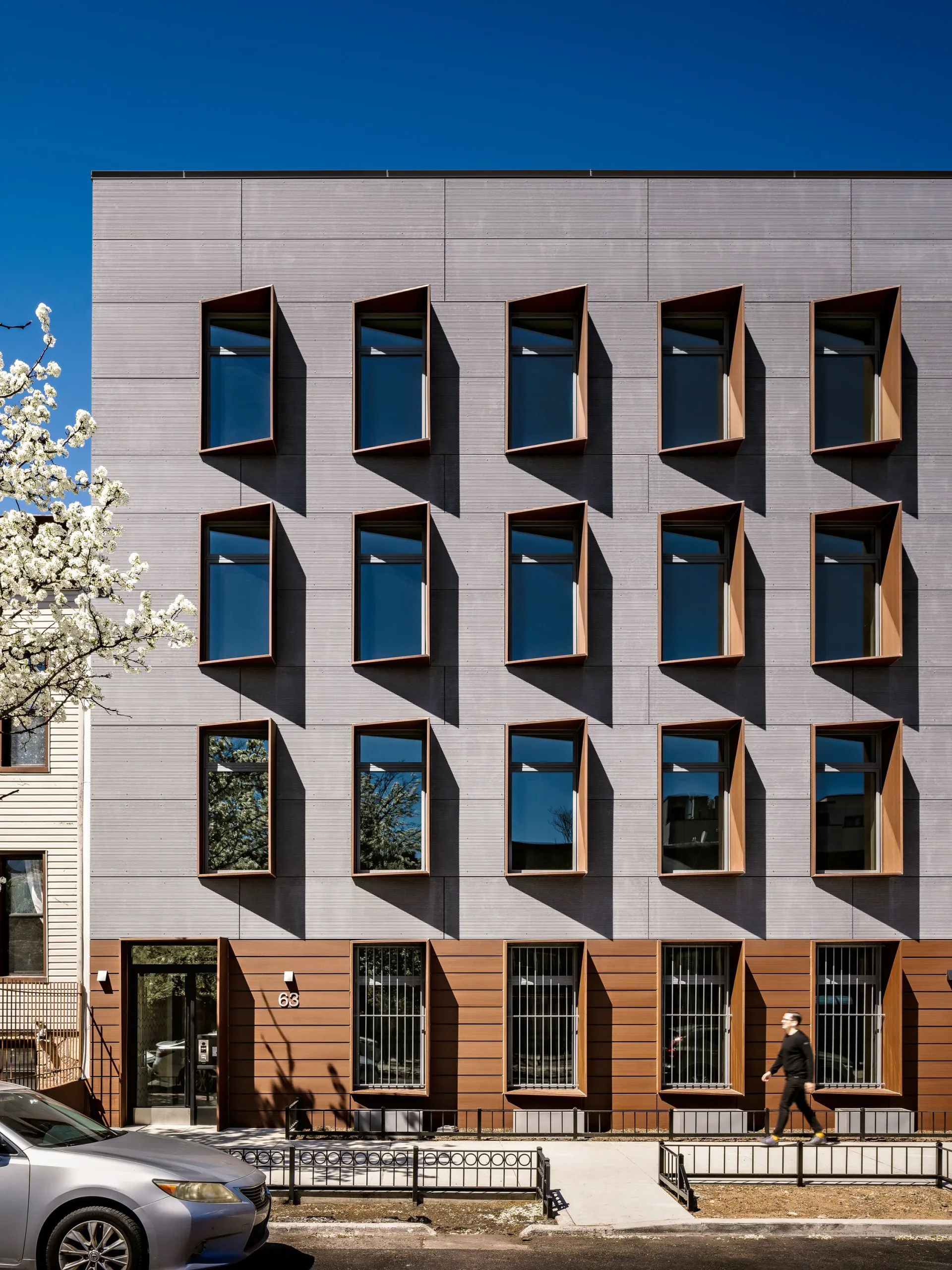 Modern residential building facade with stacked box-window modules in blue glass and warm wood cladding, white corrugated metal upper levels, designed by Suzanna Tharian.