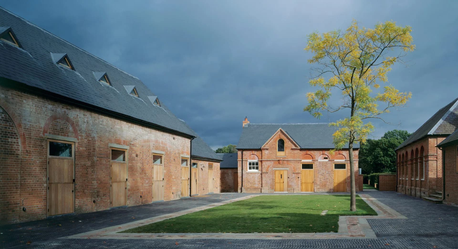 Courtyard of brick residential buildings with manicured lawn, yellow flowering tree, and dramatic stormy sky, designed by Sybille Schneider.