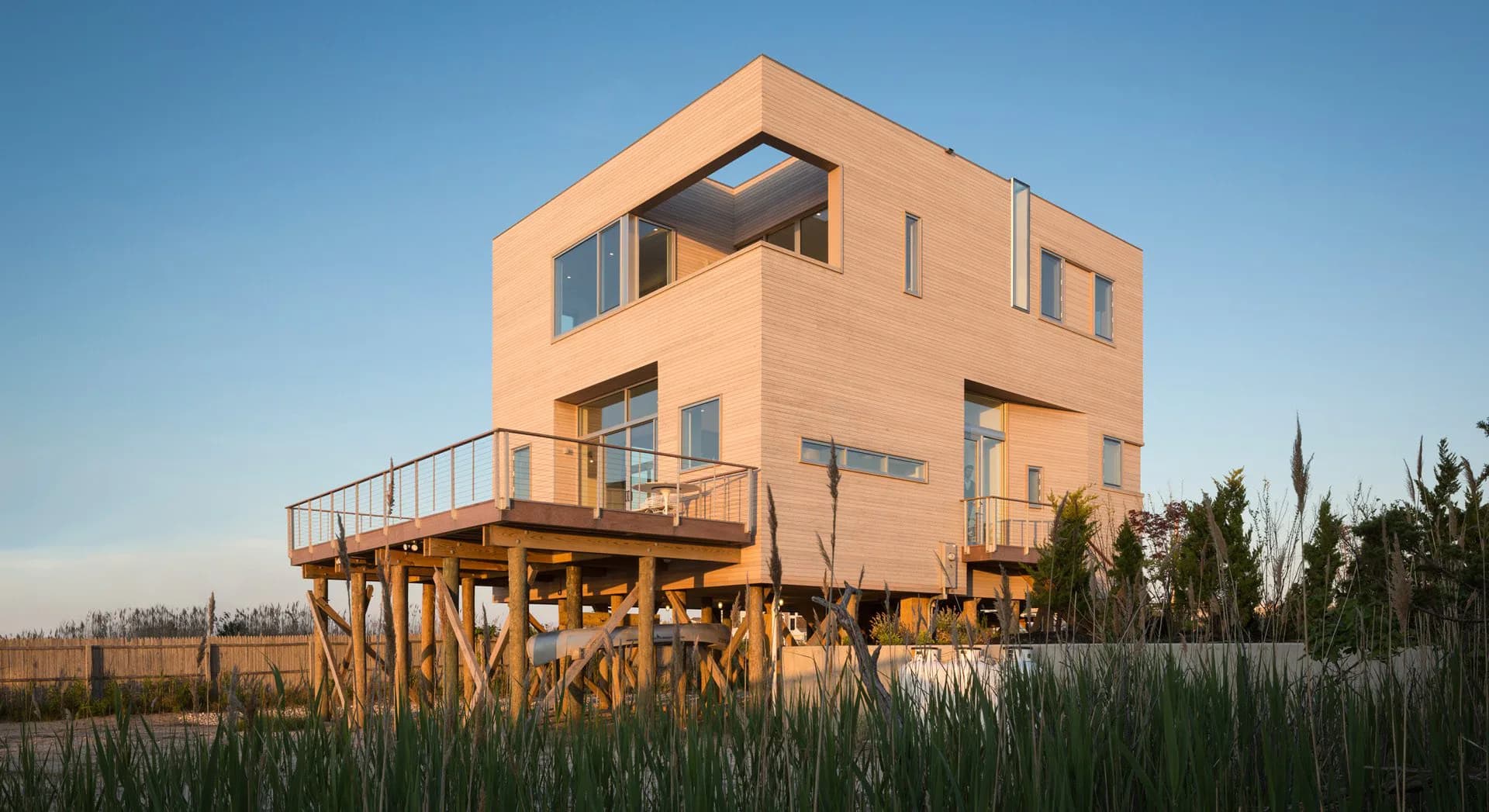 Elevated wooden house with warm peach-colored facade and horizontal window bands, set on stilts in a flat landscape with tall grasses by Sybille Schneider.