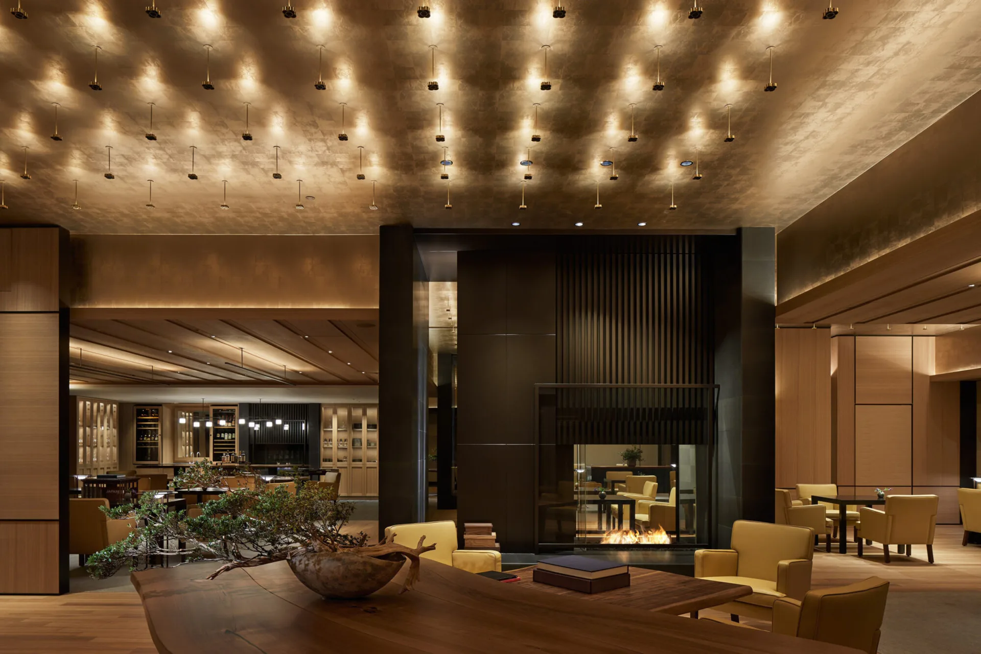 Luxury hotel lobby with warm ambient lighting, bronze and black material palette, central fireplace, and modern furnishings by Tammy Chou.