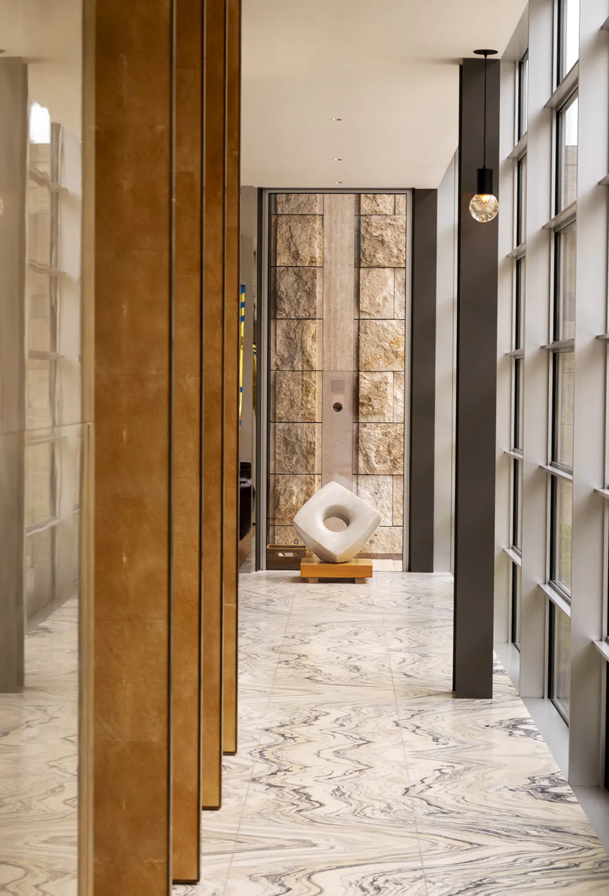 Modern marble-floored corridor with warm wood vertical elements, black framing, and sculptural white stone accent piece, designed by Tammy Chou.