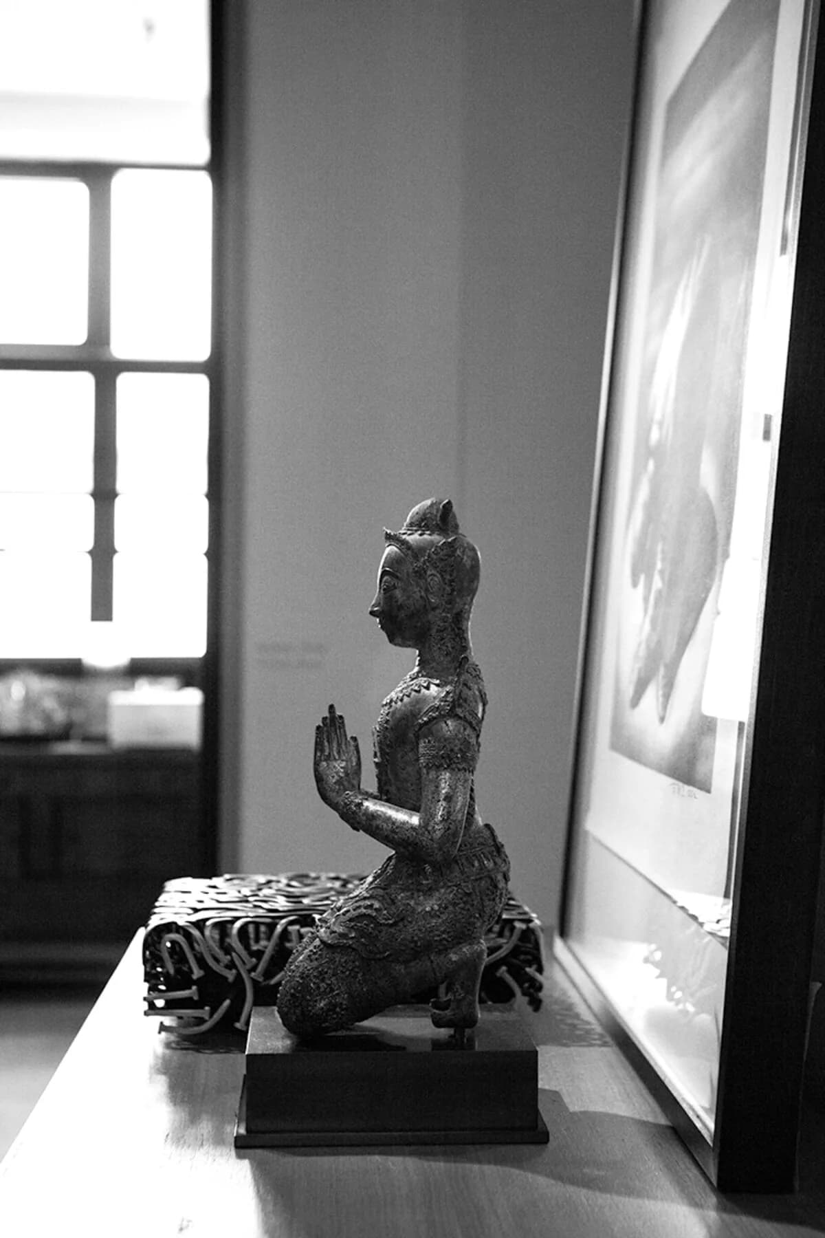 Minimalist interior displays an African carved wooden figure on a patterned textile base, positioned between a gridded window and framed artwork, designed by Tammy Chou.