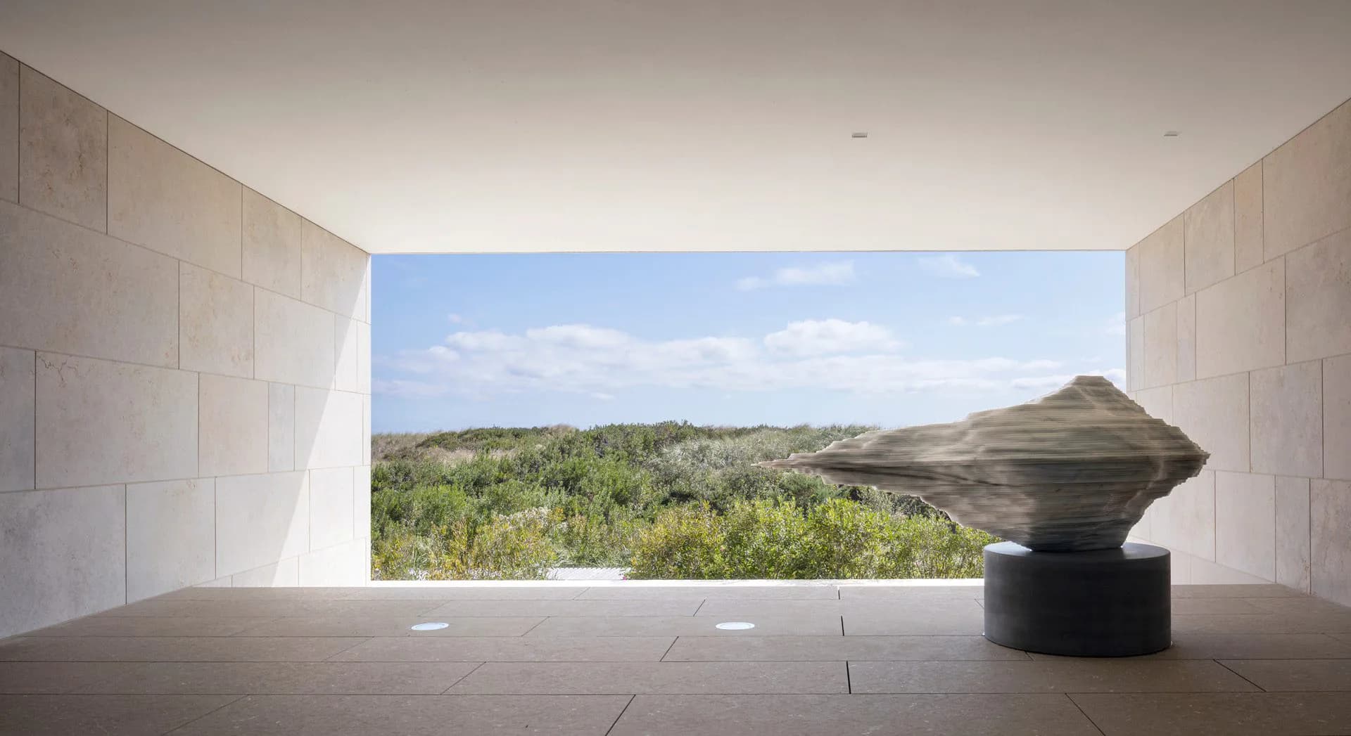 Minimalist concrete interior gallery space with dramatic floor-to-ceiling window framing desert landscape views and sculptural black vessel, designed by Tammy Mata-Lasky.