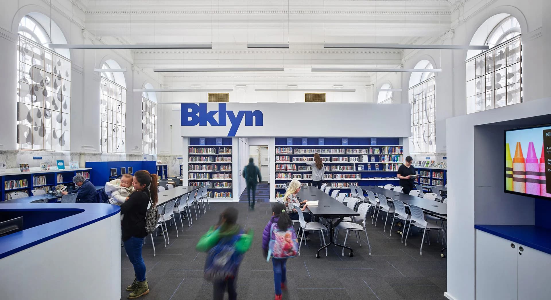 A bright, modern library interior with blue accents, white walls, natural light from arched windows, bookshelves, study tables, and large blue signage reading 'Bklyn', designed by Tammy Mata-Lasky.