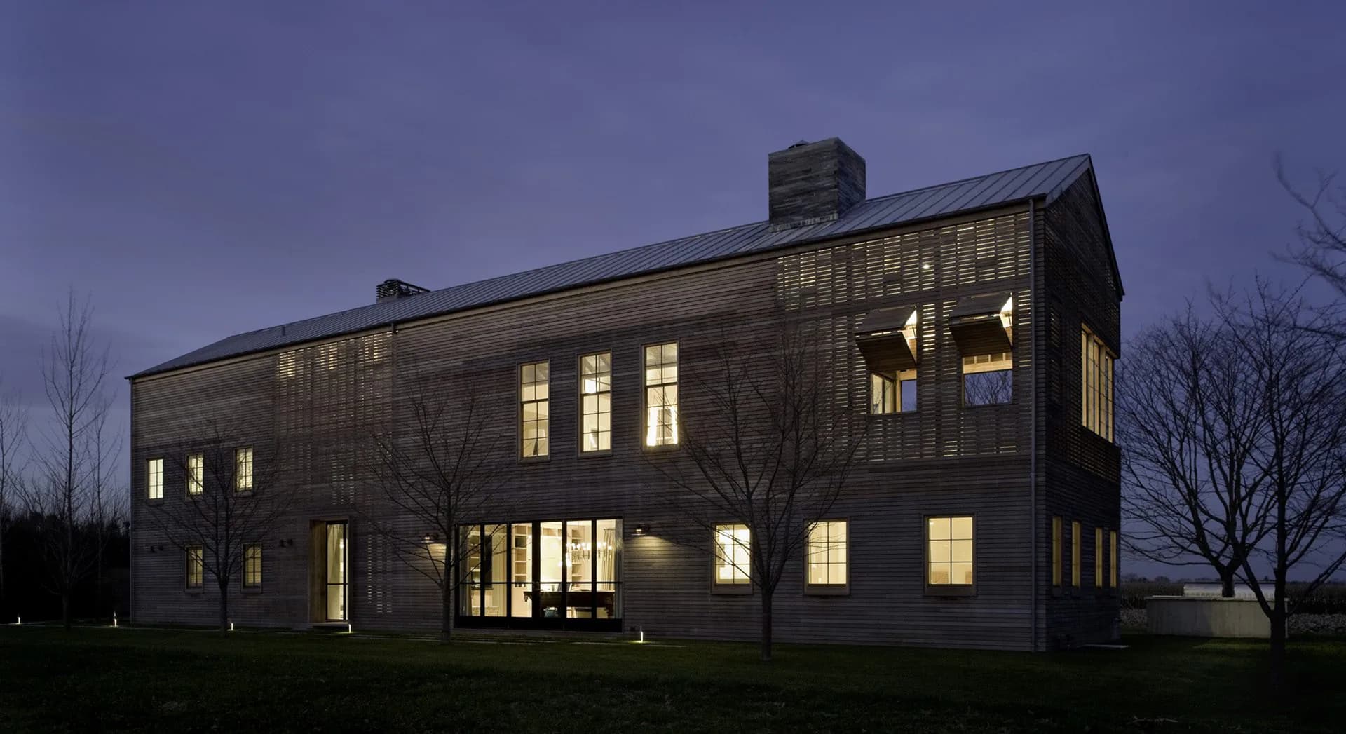 Modern residential architecture at dusk featuring a dark-clad cubic house with extensive glazing and warm interior lighting by Tammy Mata-Lasky.