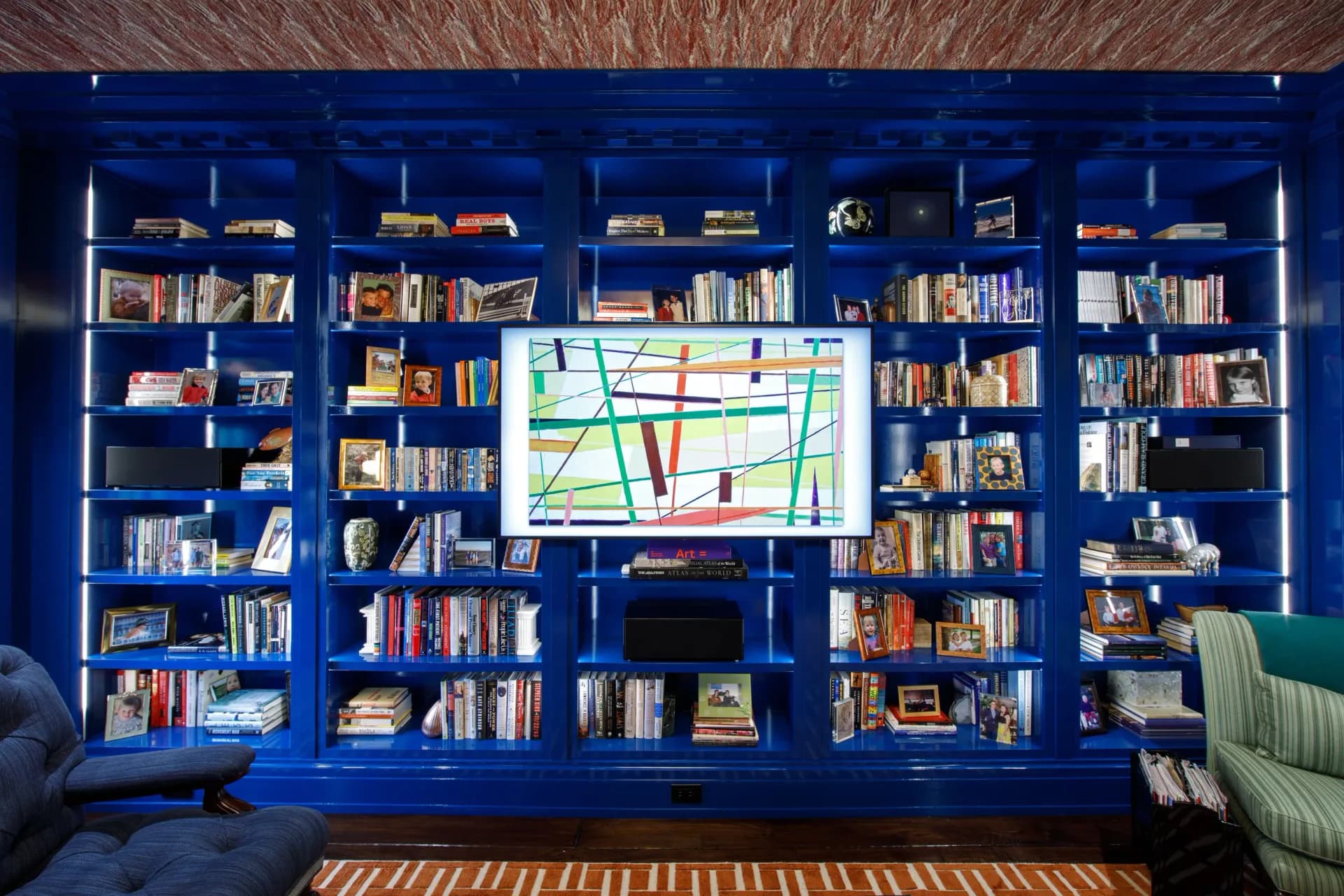 A vibrant blue built-in bookshelf wall with colorful books, a central screen displaying a map, and a striped rug, designed by Tanishkaa Kumar.