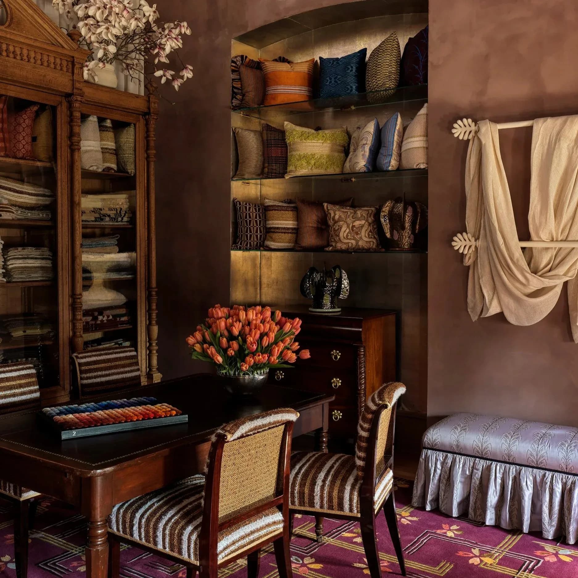 A sophisticated interior design featuring warm brown walls, wooden furnishings, decorative pillows on floating shelves, and a vibrant floral arrangement, showcasing rich textures and color coordination by Tara McCauley.