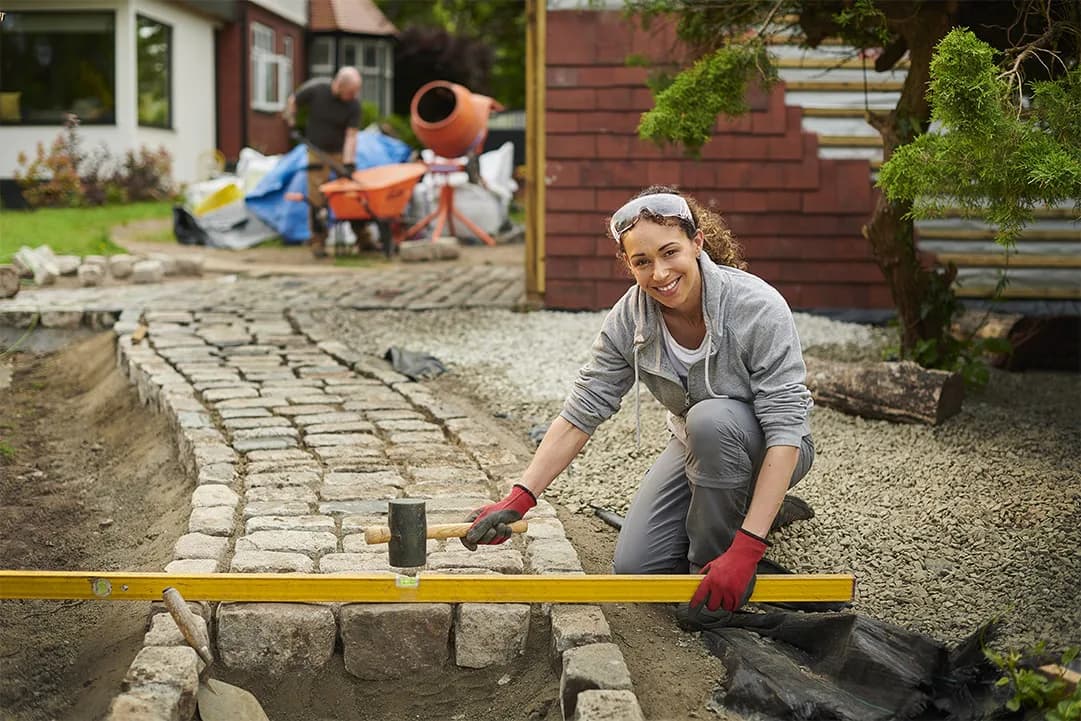 Outdoor garden patio with curved paver pathway and leveling tool in foreground, children playing in background near brick house and landscaping by Tarena Cernuto.