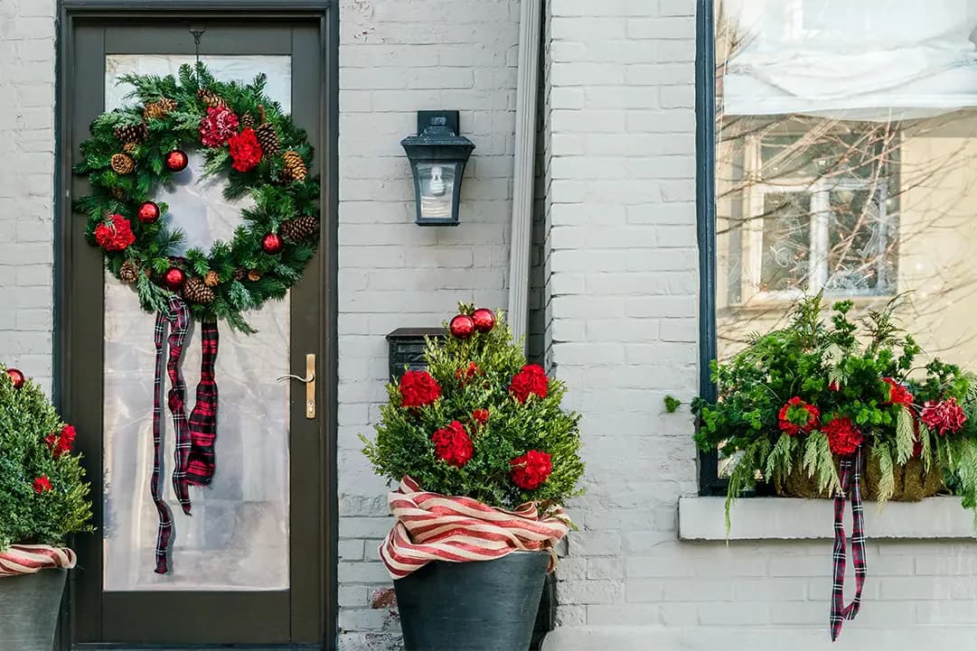White brick facade with dark-framed door and window decorated with red floral wreaths and garland by Tarena Cernuto.