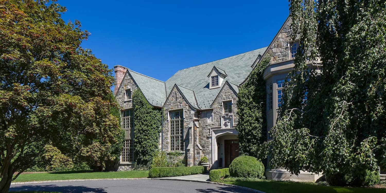 Stone Tudor-style mansion with steeply pitched roof, ivy-covered walls, arched windows, and manicured landscaping by Tasos Kokoris.