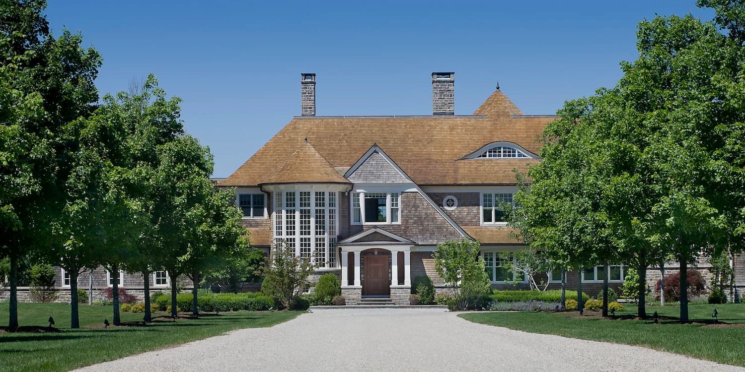 A symmetrical brick mansion with dual chimneys and a columned portico entrance, flanked by manicured trees and manicured grounds by Tasos Kokoris.
