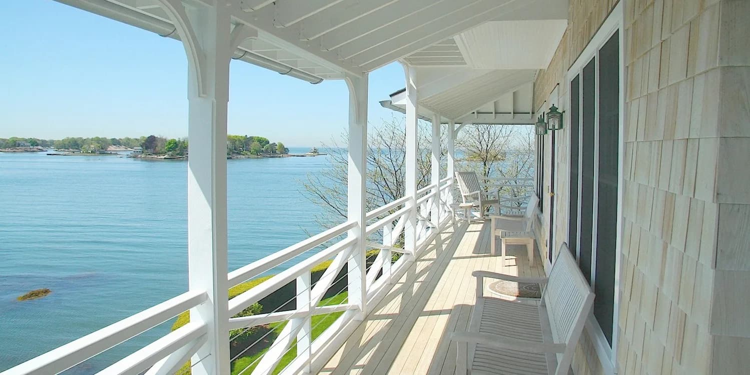 Waterfront deck with white railings and columns overlooking blue water, featuring wooden flooring and modern coastal architecture by Tasos Kokoris.