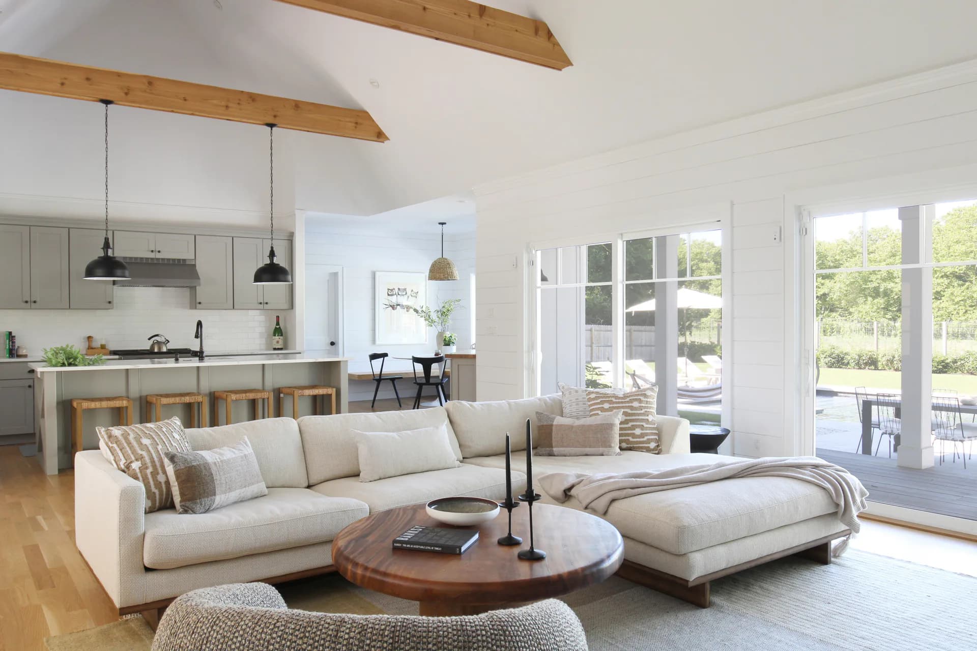 Open-plan living room with cream sectional sofa, wooden beam ceilings, kitchen with pendants visible beyond, and French doors opening to deck by Taylor Dieterich.