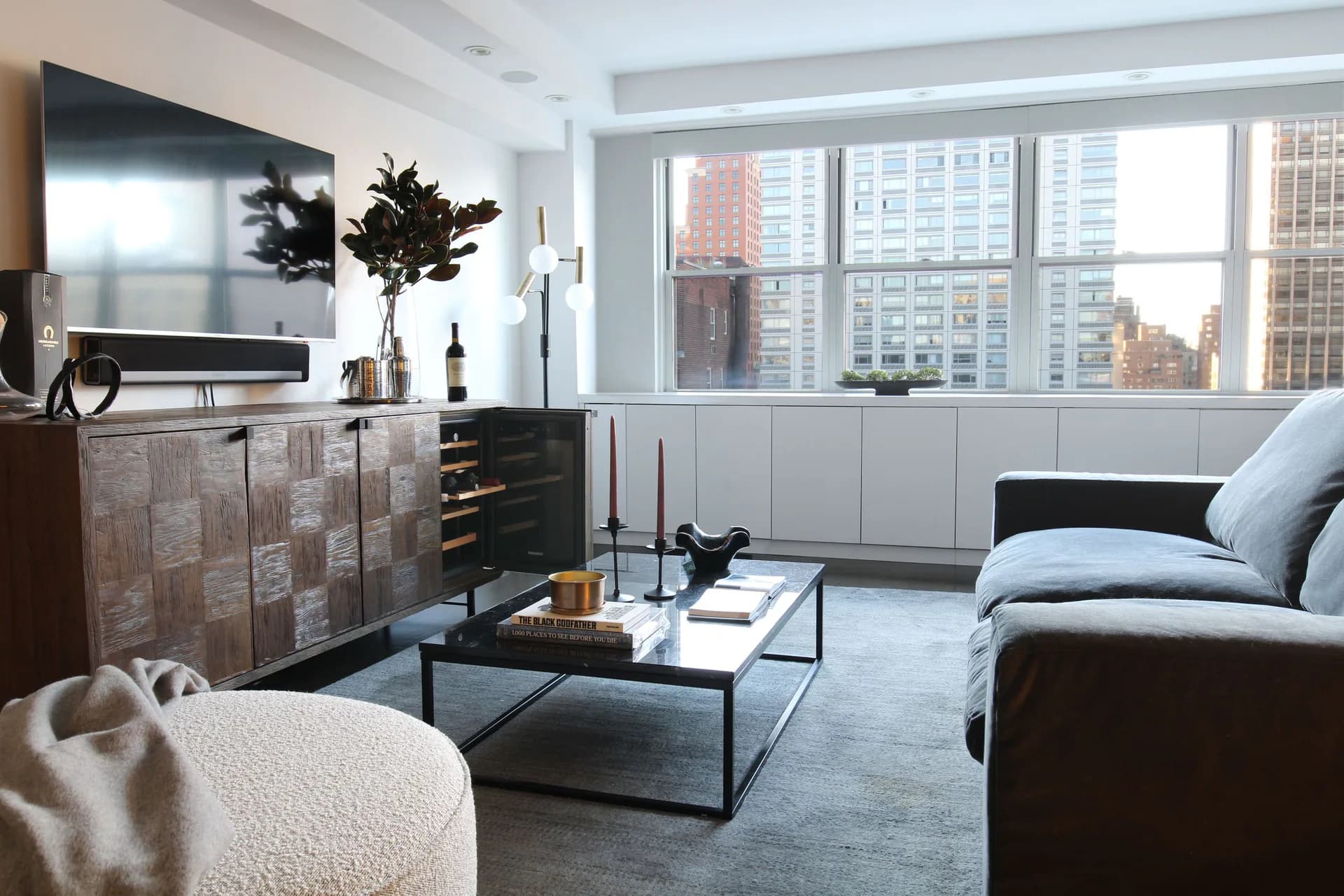 Modern living room with dark furniture, large windows overlooking cityscape, neutral palette, and wood-paneled accent wall by Taylor Dieterich.