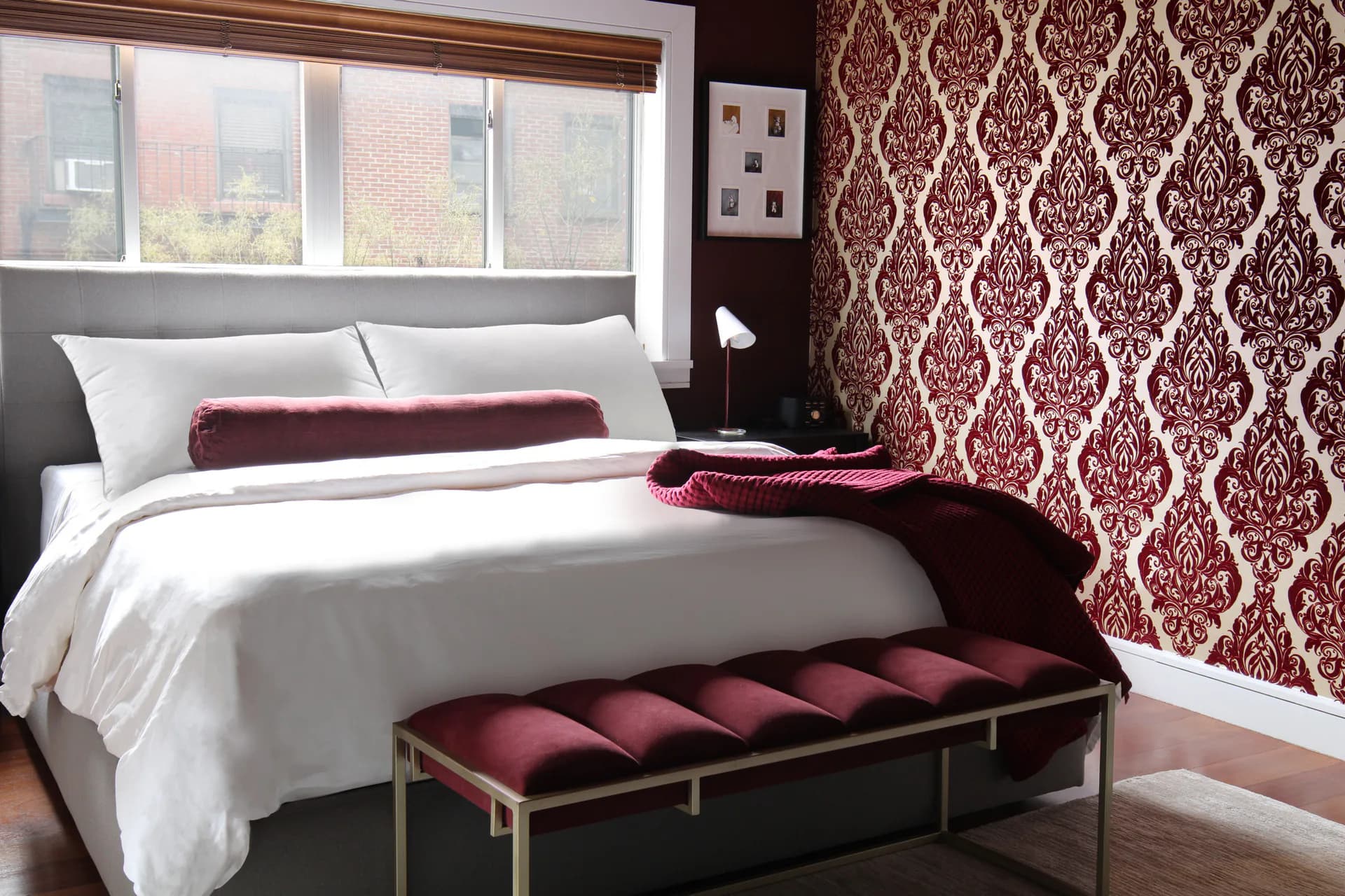 Contemporary bedroom with white bedding, burgundy damask wallpaper accent wall, upholstered bench, and large windows overlooking a city by Taylor Dieterich.