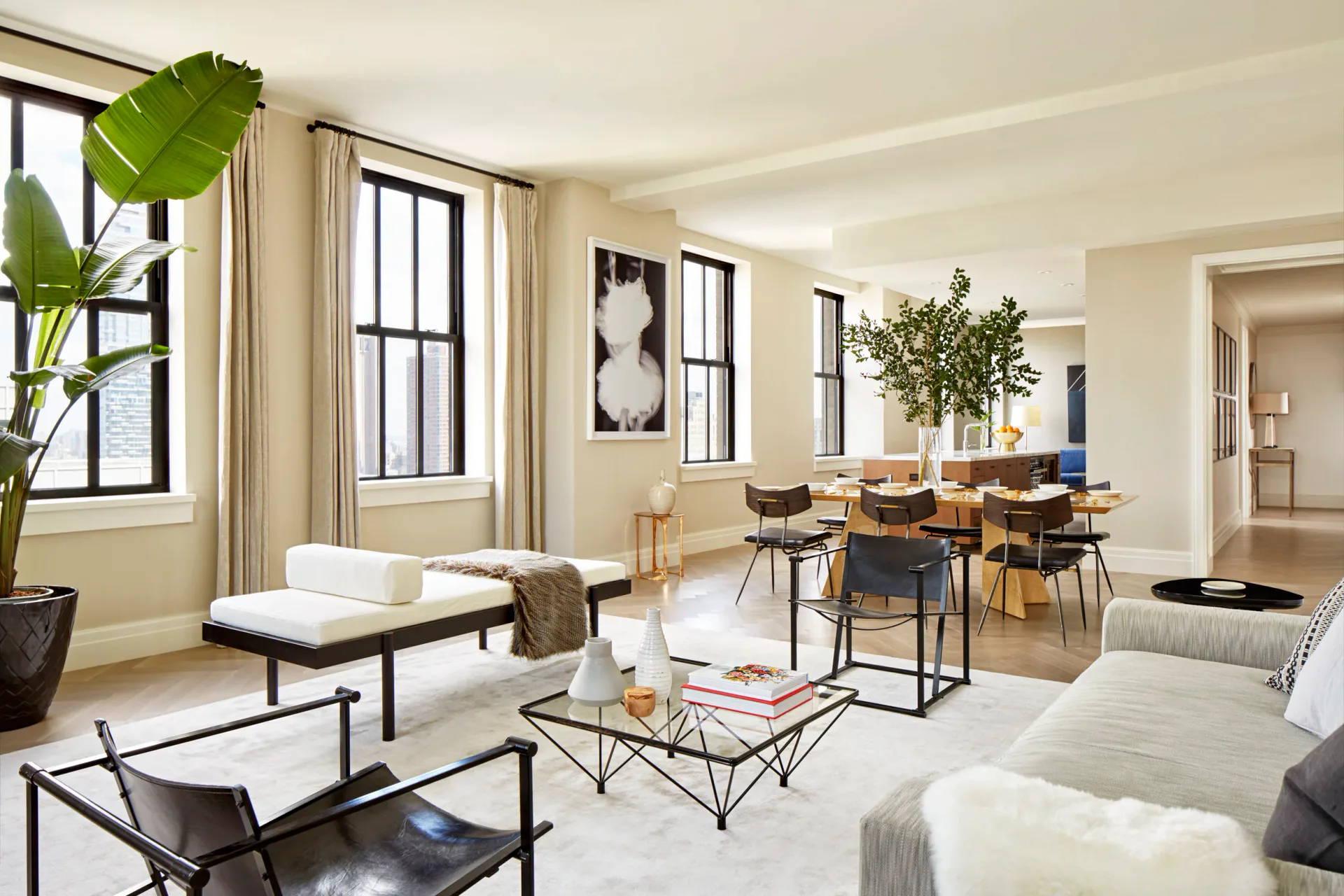 Bright open-plan living and dining space with cream walls, natural wood furniture, black-framed windows, potted plants, and modern minimalist decor by Taylor Spellman.