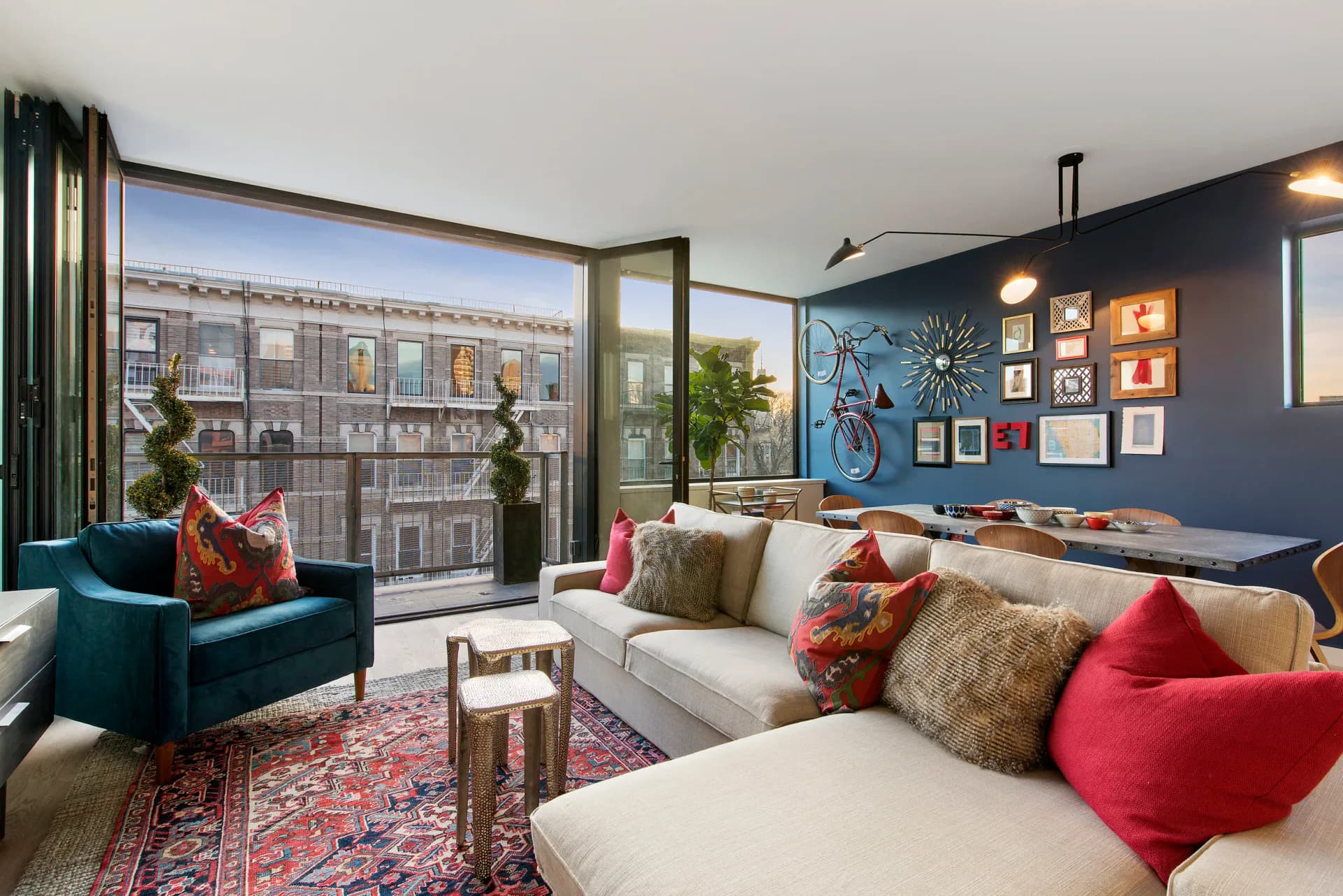 Modern living room with navy accent wall, cream sectional, teal armchair, colorful pillows, and floor-to-ceiling windows overlooking cityscape by Taylor Spellman.